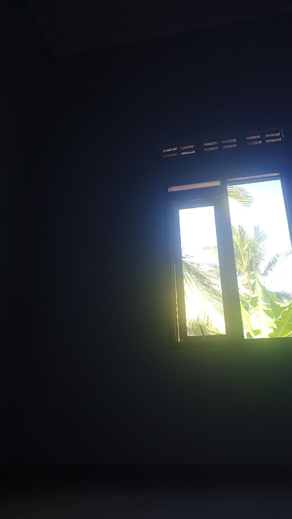 A dark room features a window with sunlight streaming through. Outside the window, palm trees and other foliage are visible. The window is framed by a wooden structure. Above the window, there are ventilation slats in the wall. The scene is set in Pakemitan, Indonesia.
