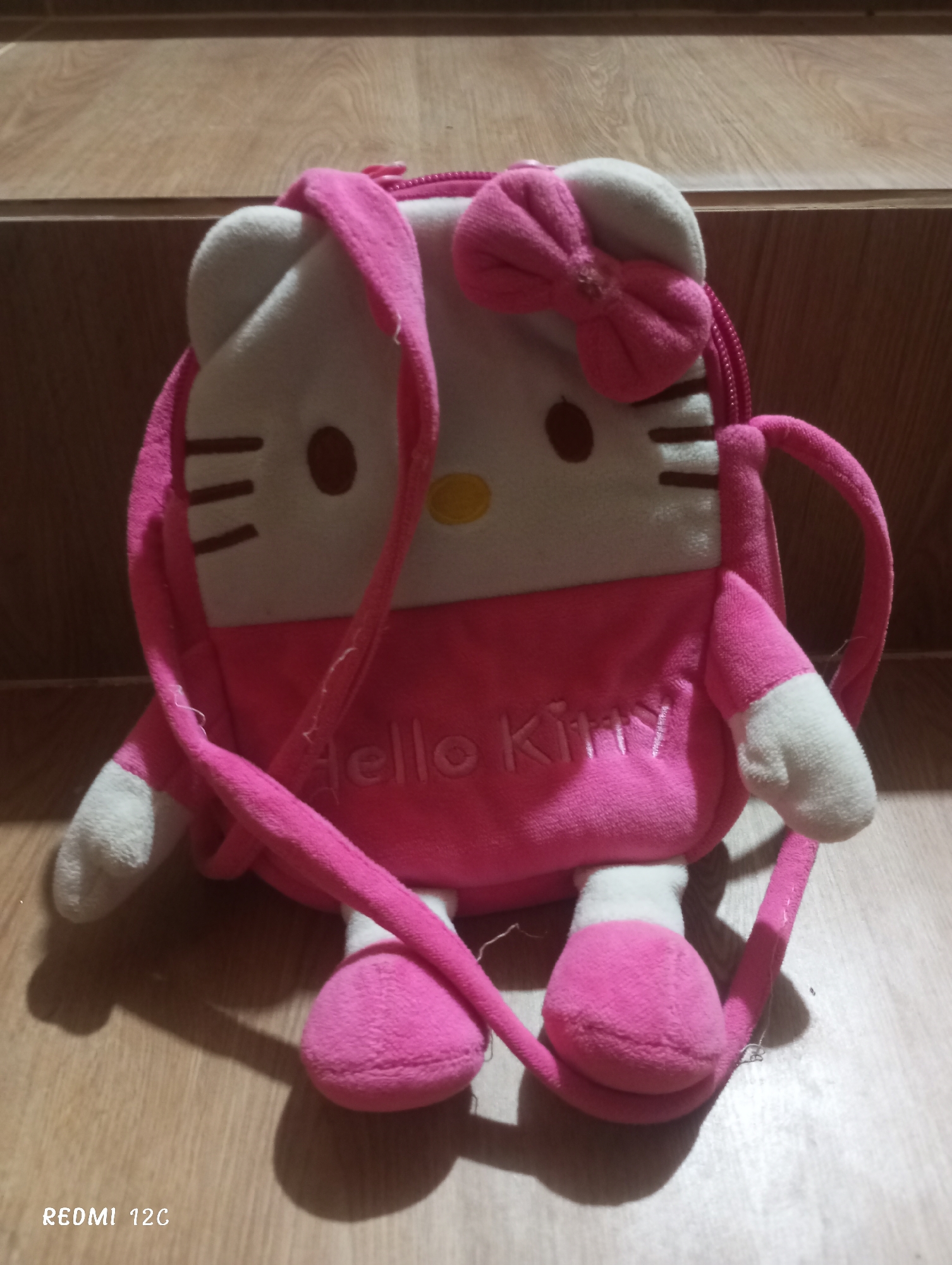 A Hello Kitty plush backpack sits on a wooden surface. The backpack is predominantly pink and white, with a pink bow on top. "Hello Kitty" is stitched across the pink midsection. The backpack has shoulder straps. The image includes a "REDI 12C" watermark in the lower-left corner. The location and details regarding the city of Lhokseumawe, Indonesia are not applicable.