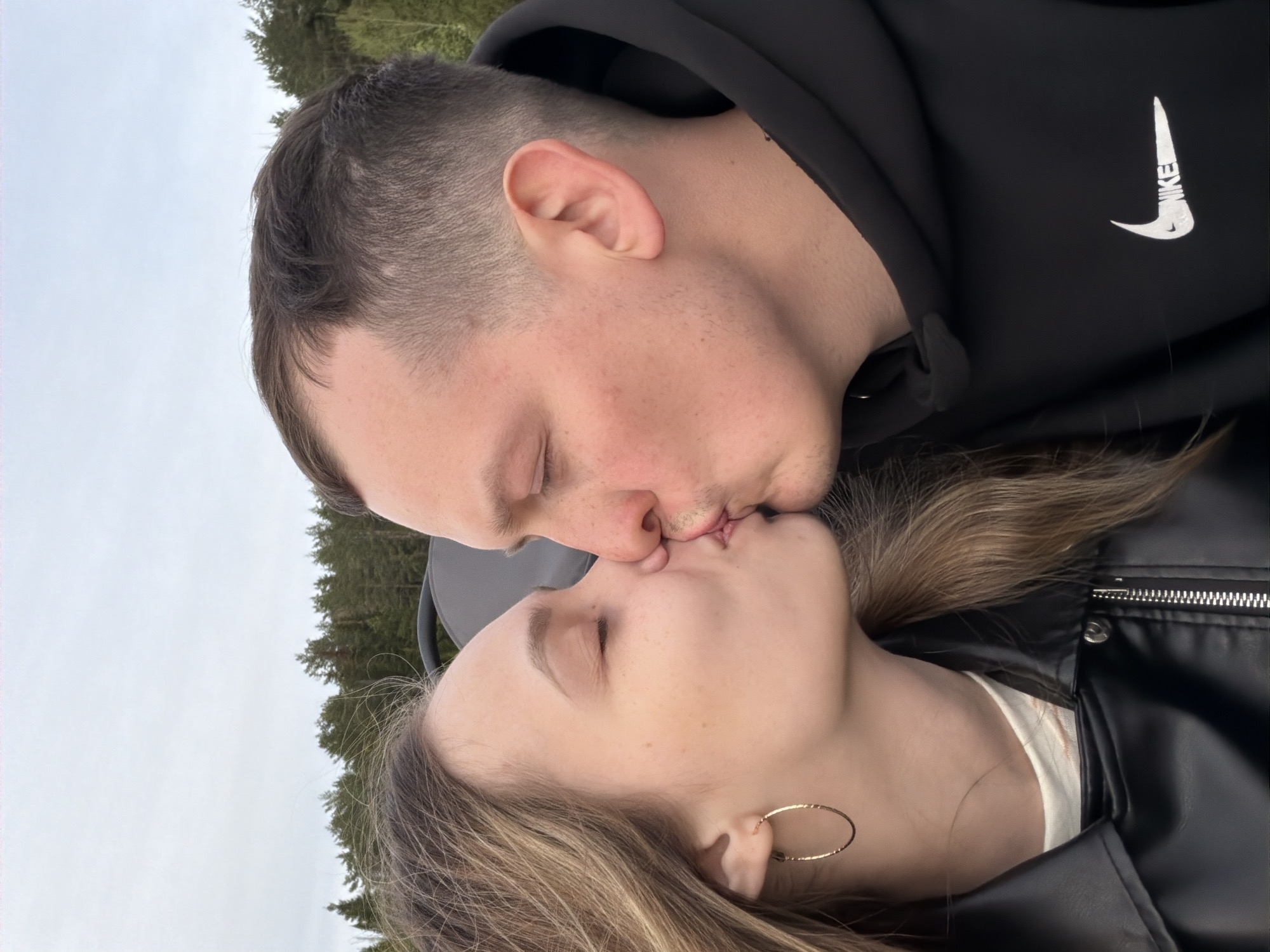 A close-up, rotated view shows a man and a woman kissing. The man has short, dark hair, and is wearing a black hooded sweatshirt with a white Nike logo. The woman has shoulder-length blonde hair and is wearing a black leather jacket over a white shirt, and a gold hoop earring. The background appears to be a sky and trees. There is no evidence suggesting the image was taken in the city of Ольховка, Russia.
