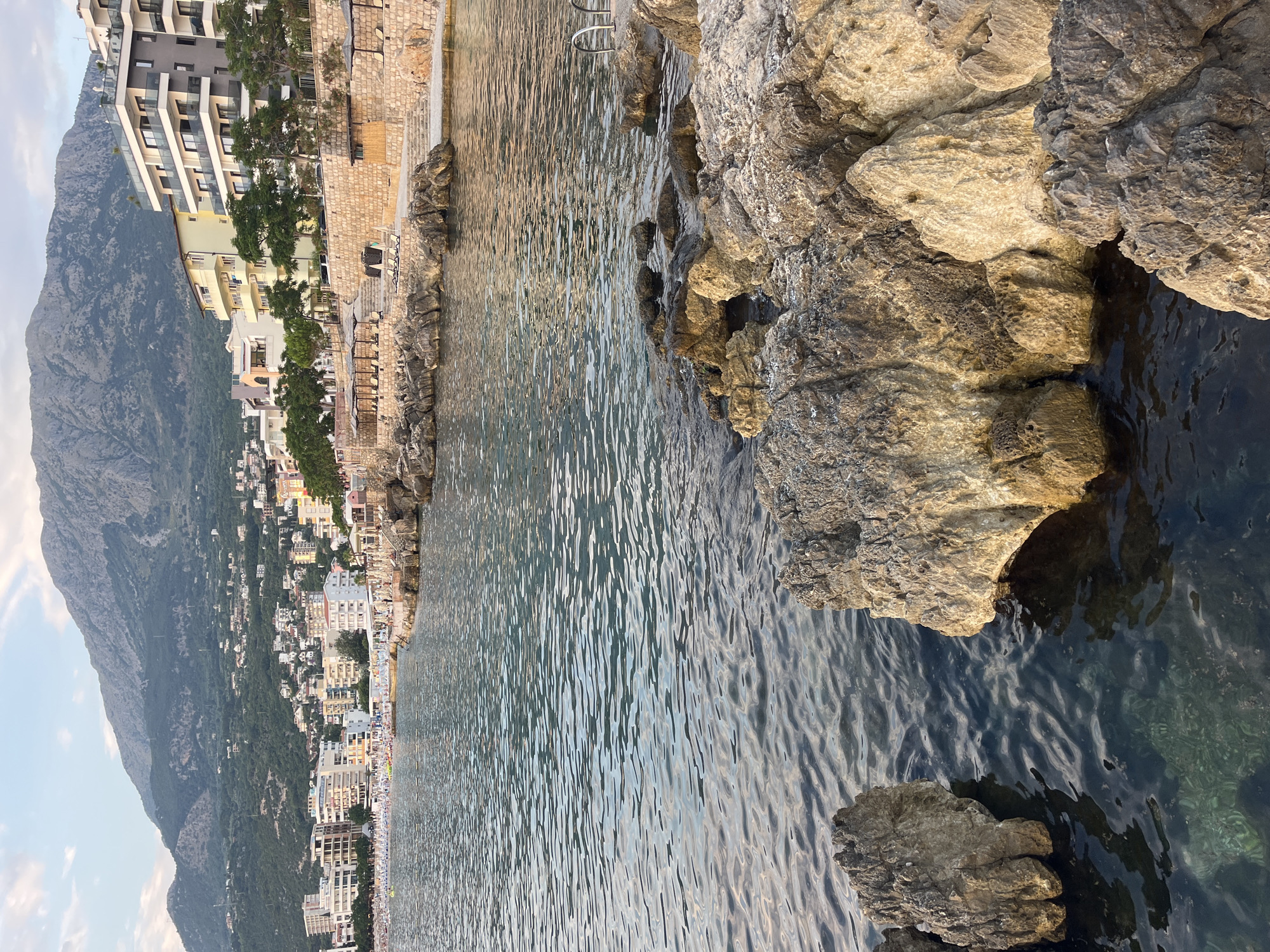 The image depicts a rocky shoreline in Pečurice, Montenegro, with a mountainous backdrop. The foreground features large, weathered rocks in the sea and on the shore. Beyond the rocks, the calm, rippling water of the sea extends towards a populated coastline. Buildings, including hotels and apartments, are clustered along the shore, and a beach with numerous sun loungers and umbrellas is visible. Trees are scattered among the buildings. The background is dominated by a large, green, and rocky mountain range. The sky is partly cloudy, with a soft, diffused light suggesting it is either early morning or late afternoon. There are no people clearly visible in the foreground or mid-ground, but the presence of beach equipment indicates activity on the beach. The overall scene is one of a coastal resort town nestled against a natural mountain landscape.