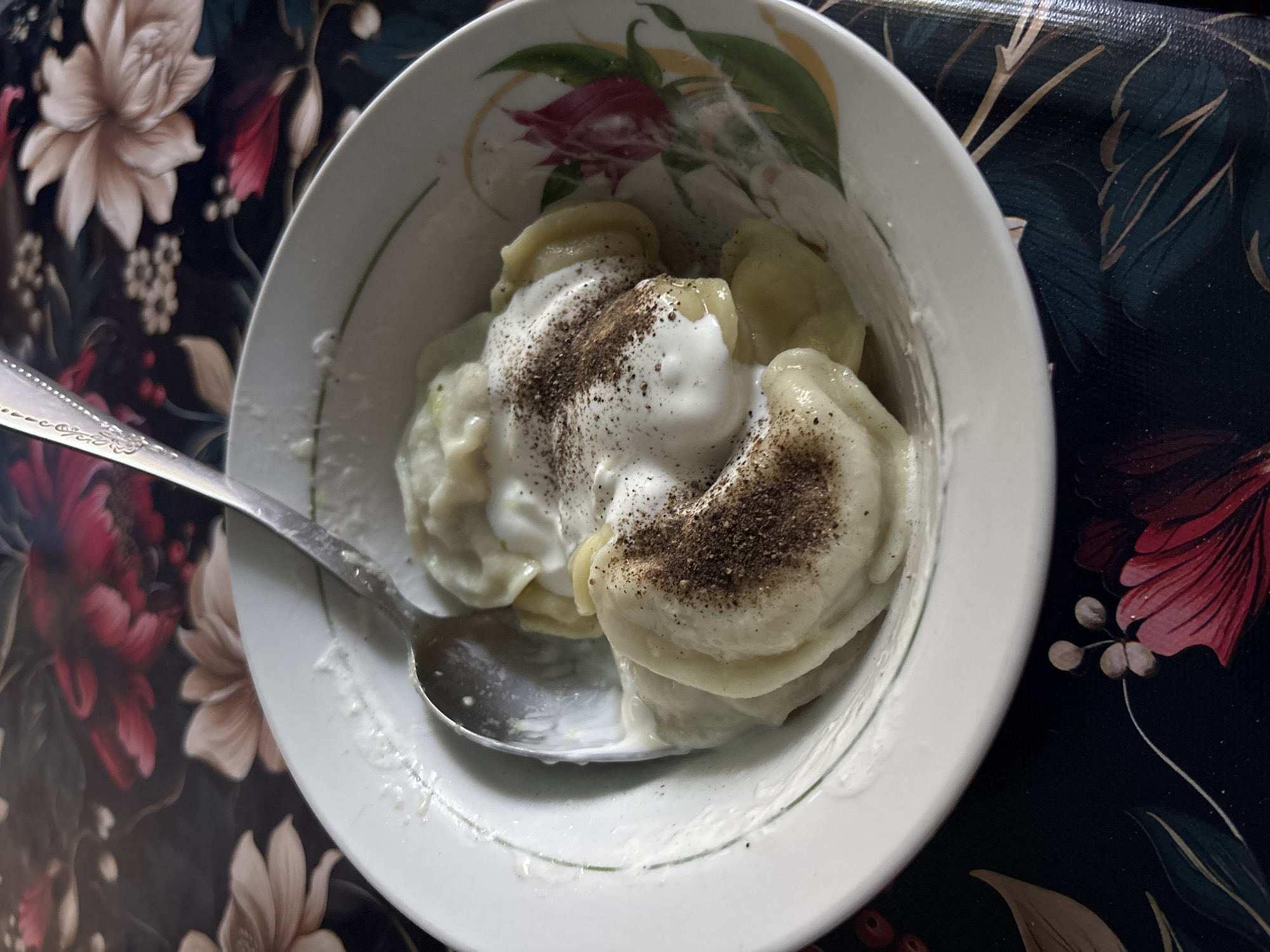 A close-up, overhead shot shows a white bowl filled with dumplings, topped with sour cream and sprinkled with pepper. A metal spoon rests in the bowl, partially submerged in the sour cream. The bowl is placed on a dark tablecloth with a floral pattern featuring large, stylized flowers in shades of pink, beige, and green. The lighting is natural, casting soft shadows on the food and the tablecloth. There are no people or animals visible in the image. The setting is a table, likely in a home or restaurant.