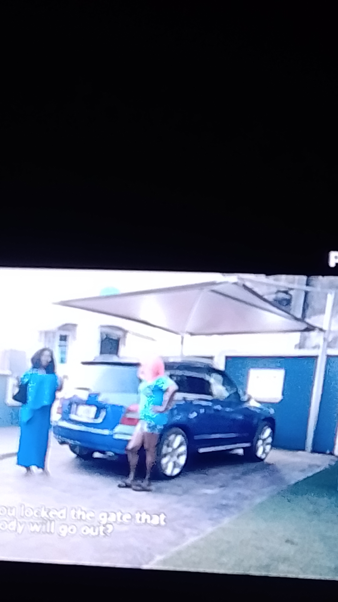 In a scene taking place in Lokoja, Nigeria, two women are standing near a blue car parked outdoors. A large, light-colored umbrella provides shade over the area. The woman on the left is wearing a long, flowing blue garment and a black bag is slung over her shoulder. The woman on the right, with pink hair, is wearing a short, patterned blue dress. She is facing away from the camera, with her hands on her hips, and is wearing sandals. The car has its trunk open. The ground is paved, and a wall or fence is visible in the background. Text superimposed on the bottom of the image reads "ou locked the gate that" and "ody will go out?".