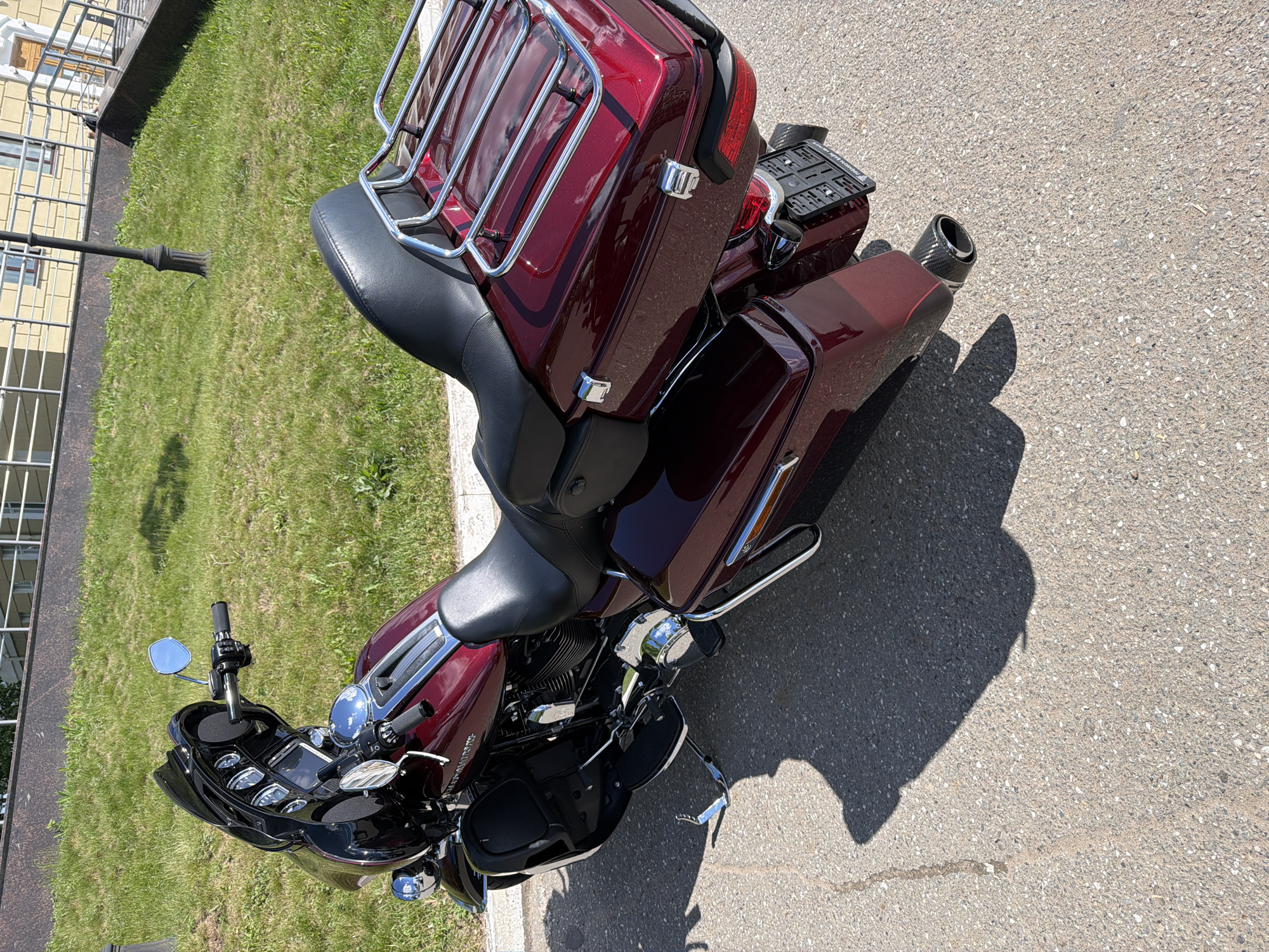 A metallic red Harley-Davidson touring motorcycle is prominently featured, parked on an asphalt surface under direct sunlight. The motorcycle displays a black "batwing" fairing with a tinted windscreen, chrome handlebars, a black two-up seat, and color-matched hard saddlebags. A chrome tubular luggage rack is mounted on the rear. The right exhaust pipe, visible at the rear, features a carbon fiber-patterned end cap. A Russian license plate, marked "RUS" at the bottom and affixed in a black holder, is attached to the rear fender. The motorcycle's chrome kickstand is deployed, casting a distinct shadow on the pavement, alongside the shadow of a lamppost.
Behind the motorcycle, a well-maintained green grass slope ascends. This slope is bordered by a dark retaining wall that separates it from a light-colored, multi-story building with windows and white railings in the background. A decorative black lamppost stands on the grass to the right of the motorcycle. Further to the right, a set of stairs with railings leads to an entrance of the background building. No people are present in the scene. The setting is located in the city of Votkinsk, Russia.