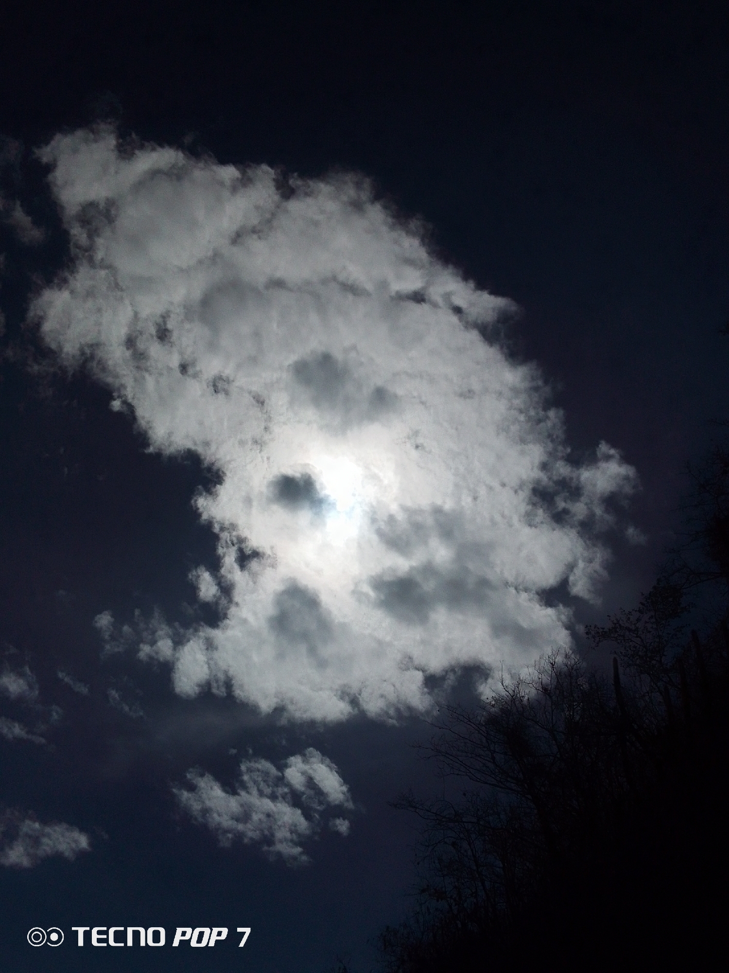 The image shows a cloudy sky at Pedro González, Venezuela. A large cloud dominates the center, with a brighter area possibly indicating the sun shining through. The sky is otherwise dark. In the lower right corner, there are some dark silhouettes of trees. The bottom left corner contains the text "TECNO POP 7".