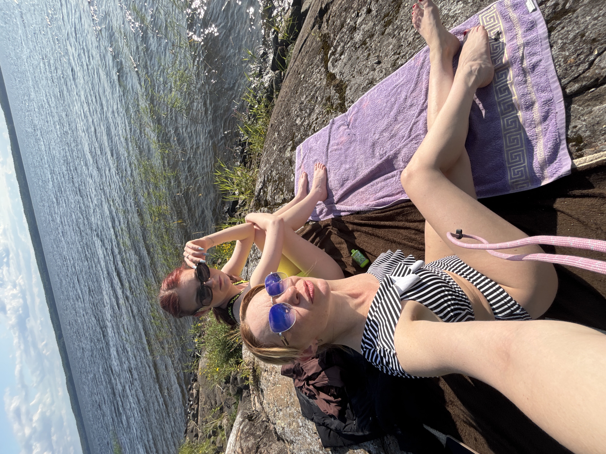 Two women are sunbathing on rocks by a lake. The woman in the foreground is wearing a black and white striped bikini and blue sunglasses. The woman behind her is wearing a yellow bikini and dark sunglasses. Both women are looking at the camera. The background shows a lake with trees on the far shore, under a partly cloudy sky. The scene appears to be in the vicinity of Shuiskaya Chupa, Russia, given the natural landscape.