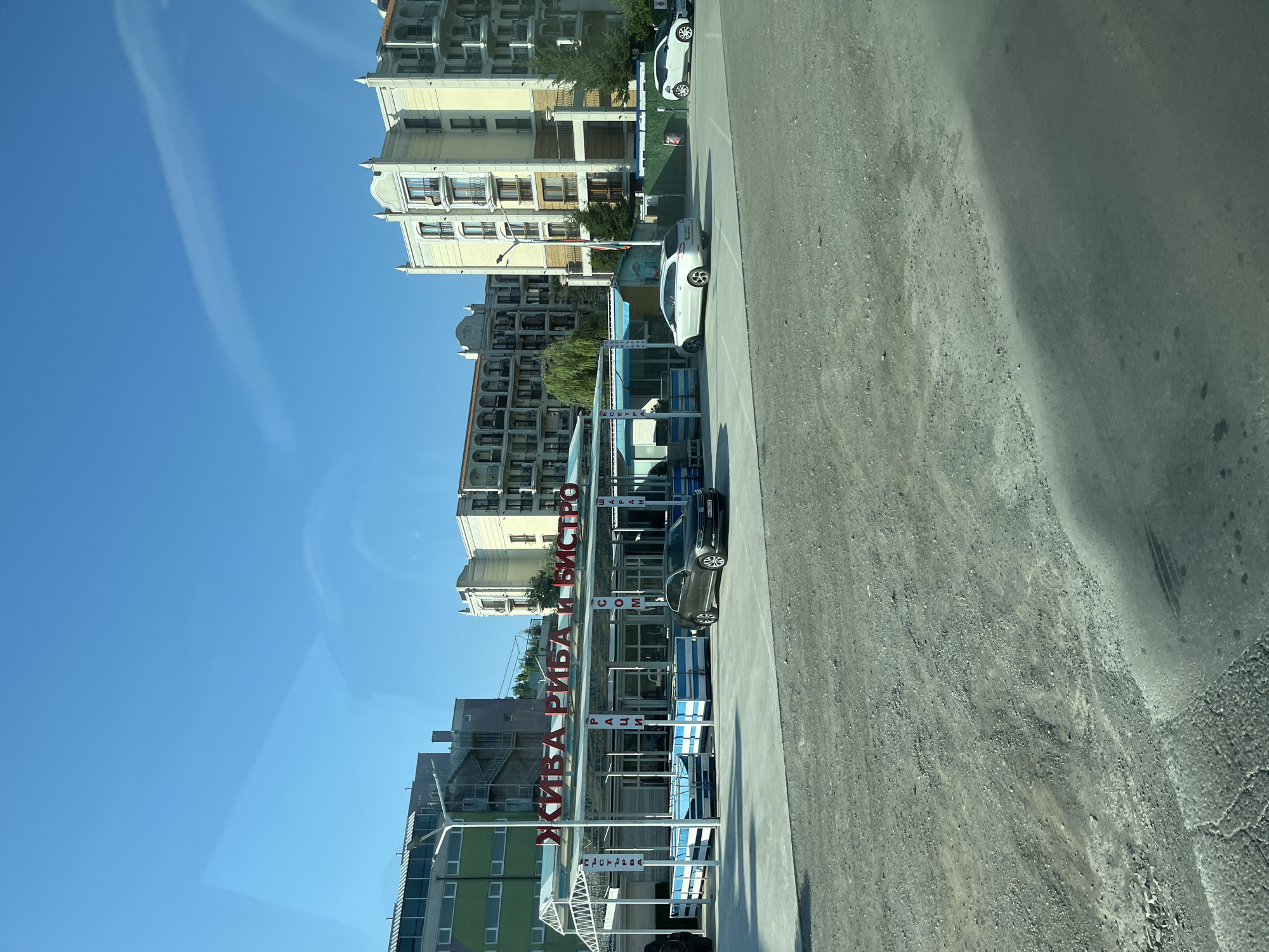 This image shows a street view of a building with a sign in Cyrillic script that reads "ЖИВА РИБА И БИСТРО," which translates to "LIVE FISH AND BISTRO." The building appears to be a restaurant or market. There are several cars parked in front of the establishment. In the background, there are multi-story apartment buildings. The sky is clear and blue. The overall scene suggests a commercial area in Nesebar, Bulgaria, likely a coastal town given the presence of a fish establishment.
