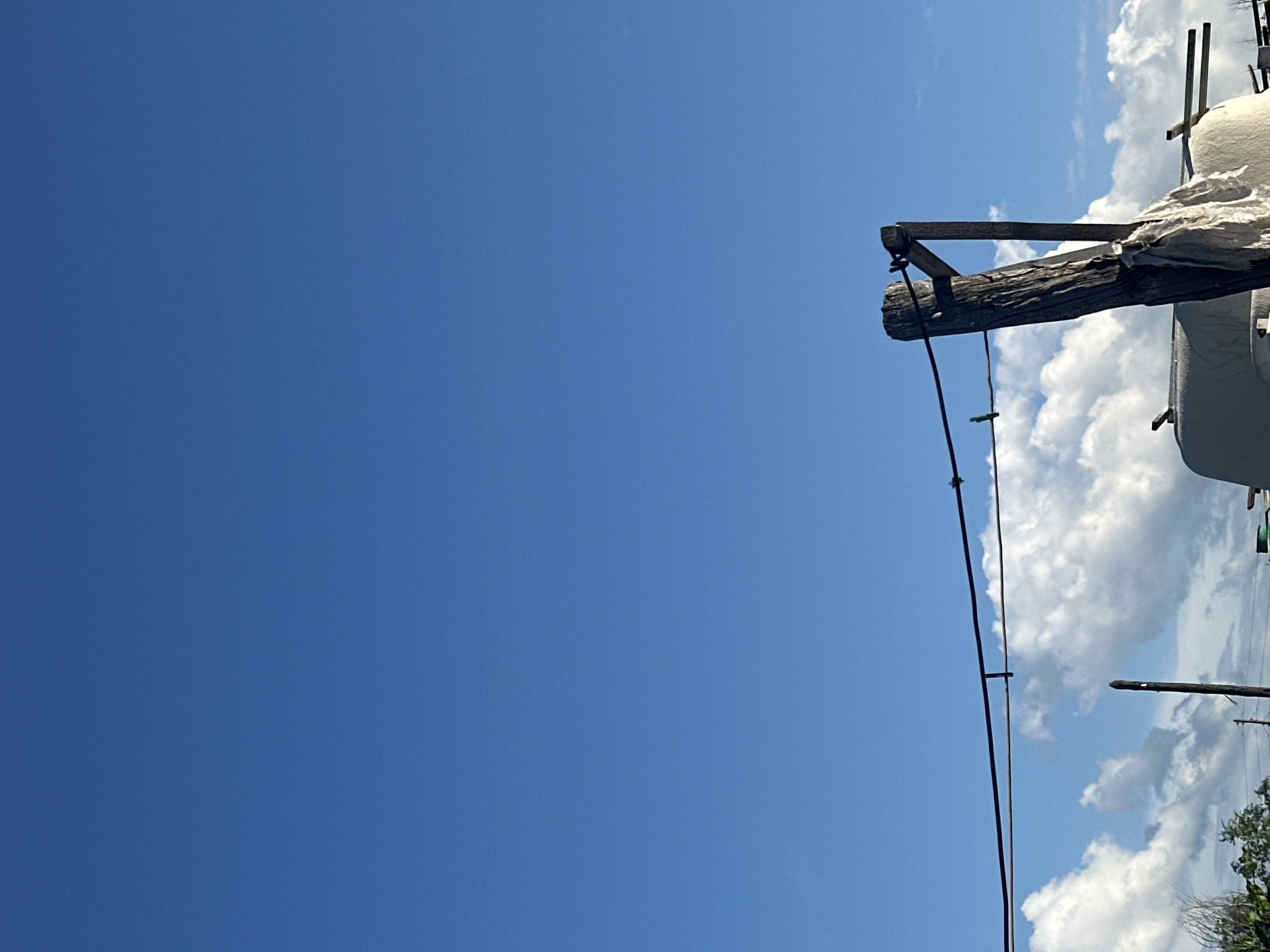 This media file shows a close-up, low-angle shot of a wooden pole against a clear blue sky and some fluffy white clouds. The pole appears weathered and rough, with a diagonal wooden brace attached to it. A dark wire runs down from the pole, with a small green clothespin attached to it. In the background, to the right of the pole, is a white, possibly tarp-covered object supported by a wooden structure, and some greenery is visible at the very bottom right. The scene suggests an outdoor, possibly rural setting in Tobyl, Kazakhstan, on a sunny day with fair weather. The composition focuses on the texture of the wood and the vastness of the sky. There is no visible text in the image.