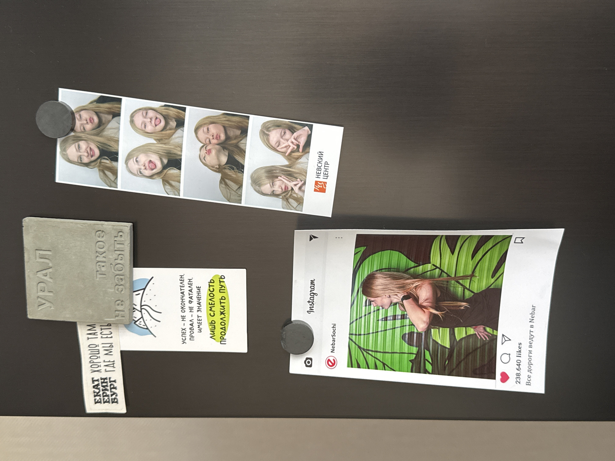 The image shows a dark, potentially wooden, surface (likely a refrigerator door or cabinet) affixed with a set of magnets holding various papers and ephemera. A strip of photo booth pictures featuring a young woman with blonde hair in various playful poses is attached with a magnet on the left. Next to that is a concrete souvenir labeled "УРАЛ" (Ural) with the phrase "такое Не забыть" (something not to be forgotten). Below this is a small rectangular paper saying, "ЕКАТ ХОРОШО ТАМ БУРГ ГДЕ МЫ ЕСТЬ ЕРИН," a reference to the city of Yekaterinburg. Beside this, a paper has a quote that reads, "УСПЕХ - НЕ ОКОНЧАТЕЛЕН, ПРОВАЛ – НЕ ФАТАЛЕН. ИМЕЕТ ЗНАЧЕНИЕ ЛИШЬ СМЕЛОСТЬ ПРОДОЛЖИТЬ ПУТЬ." (Success is not final, failure is not fatal. It is the courage to continue that counts.) Another magnet is holding a printout of an Instagram post from "NebarSochi" showing a woman in front of a green patterned backdrop, with the caption "238.640 likes Все дороги ведут в Nebar" (All roads lead to Nebar). A logo for "Невский центр" is also visible on the photo booth strip. No direct location in Perm, Russia, is provided, though it is in the broader Ural Region.