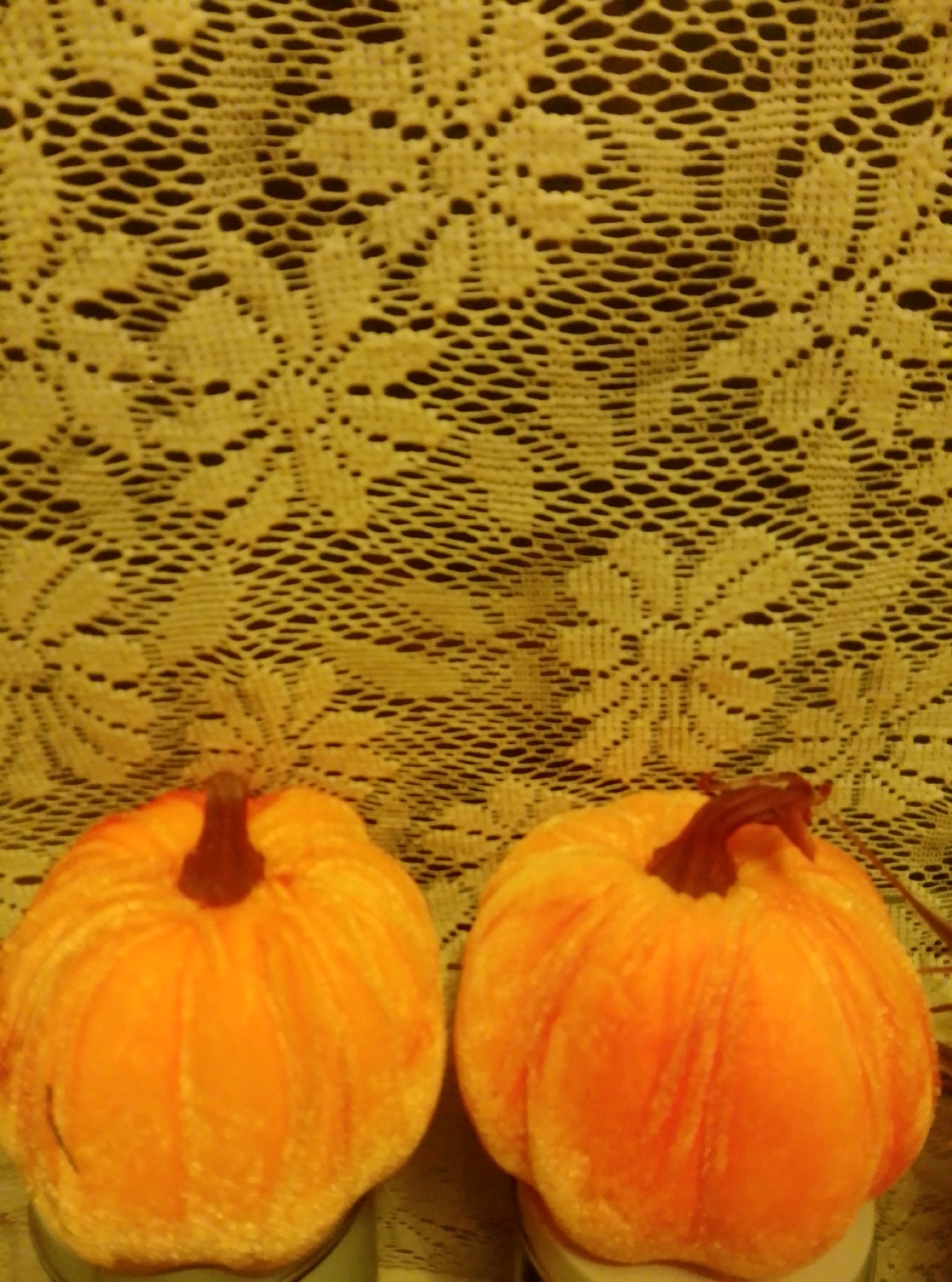The image depicts two artificial pumpkins against a backdrop of yellow lace. The pumpkins are orange, with visible stem details and ribbed surfaces. One pumpkin is positioned on the left side of the frame, and the other is to its right, slightly further back. The lace has an intricate, floral pattern with a dark background showing through the openwork. The scene is indoors, with the lighting casting a warm glow. There is no indication of the city of Wedel, Germany within the image.