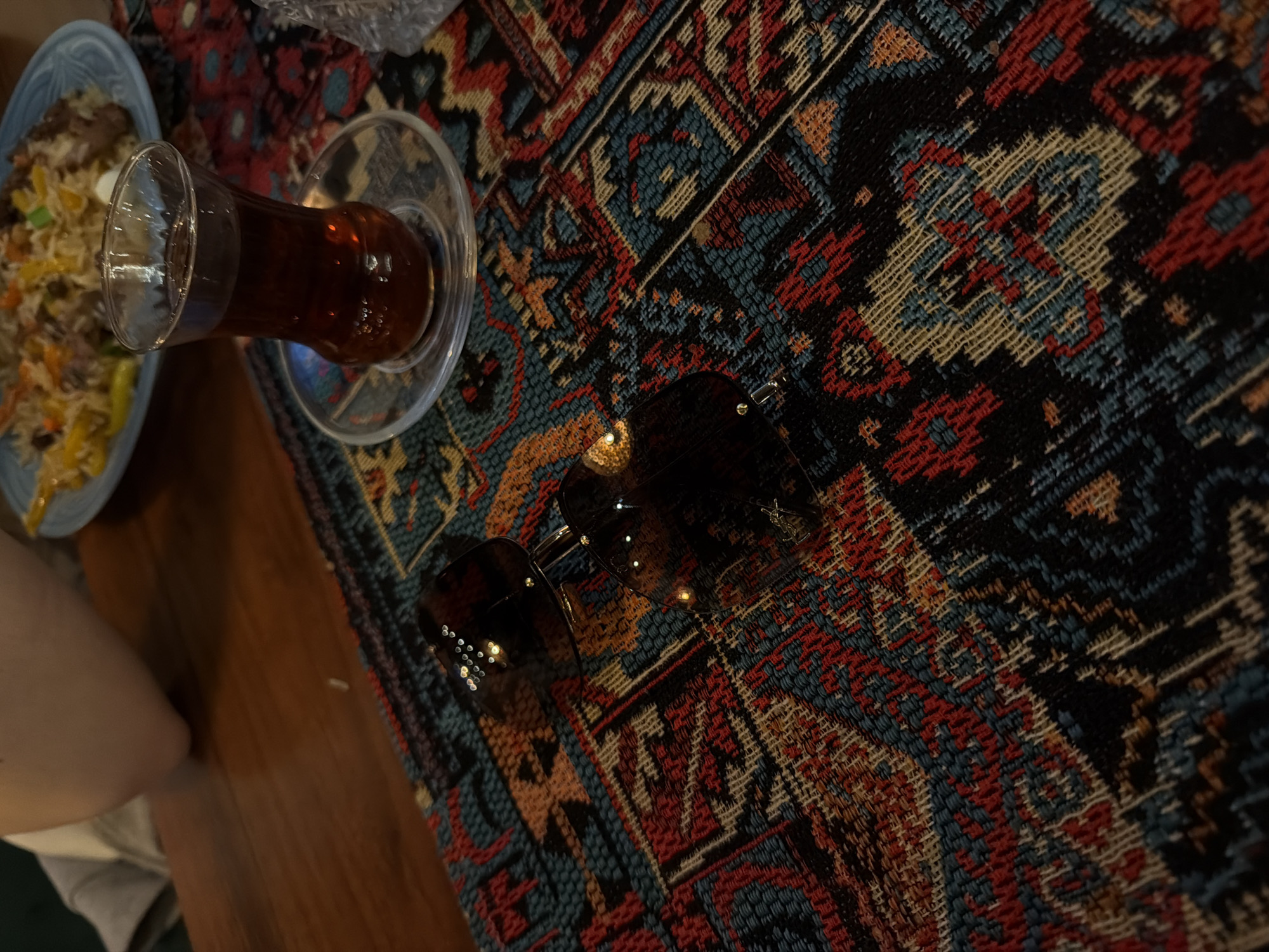 The image shows a still life arrangement on what appears to be a patterned rug, possibly of Persian or Middle Eastern design. A partially consumed plate of food is visible, containing a yellow-toned dish with visible ingredients. A traditional glass of tea, on a saucer, sits adjacent to the plate. A pair of sunglasses rests on the rug, slightly obscuring the pattern. The rug itself features intricate designs in shades of red, blue, beige, and black. Part of a person's arm and clothing is visible in the lower left corner of the frame. The overall impression is of a casual setting. No specific location in Warsaw, Poland is evident from the image alone.