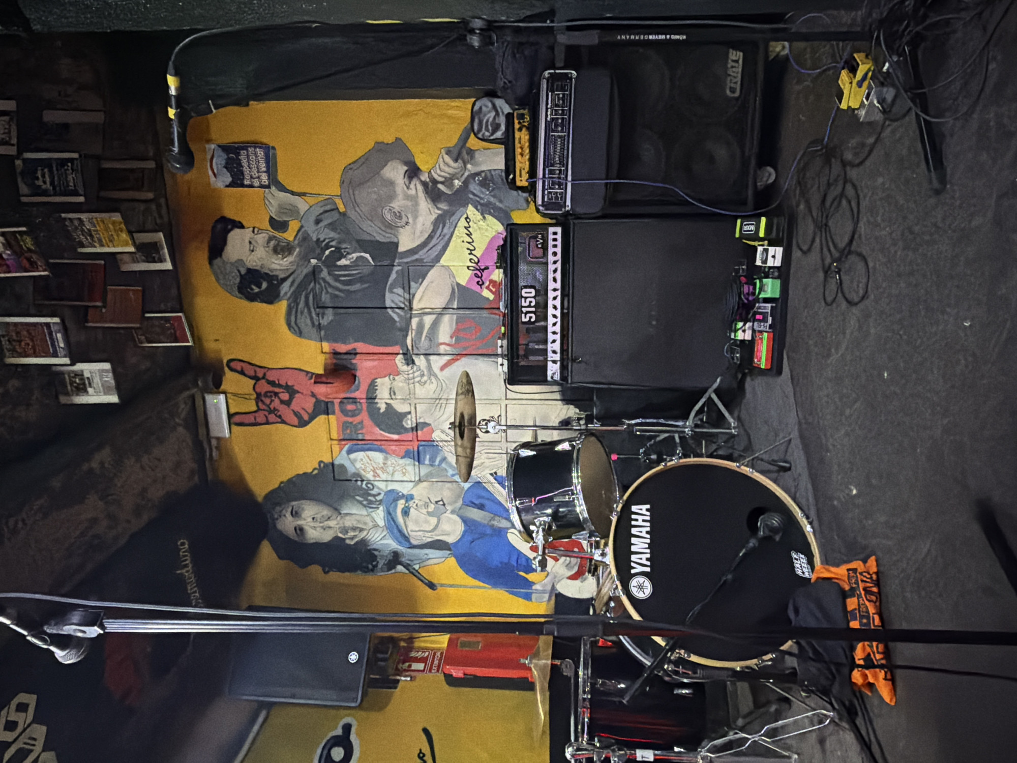 The image shows a music stage. Equipment visible includes a Yamaha drum set, guitar amplifiers (Crate, 5150 EVH), a pedalboard, and microphones. There is a colorful mural on the wall behind the stage featuring portraits of people and the words "ceferino", "remoduro", and "Respeta el descanso del veinat". Small posters or album covers are affixed to a wall on the left side of the image. An orange towel or cloth with writing on it rests against the drum set. A red fire extinguisher can be seen behind the drums. The mural's content points towards Barcelona, Spain.