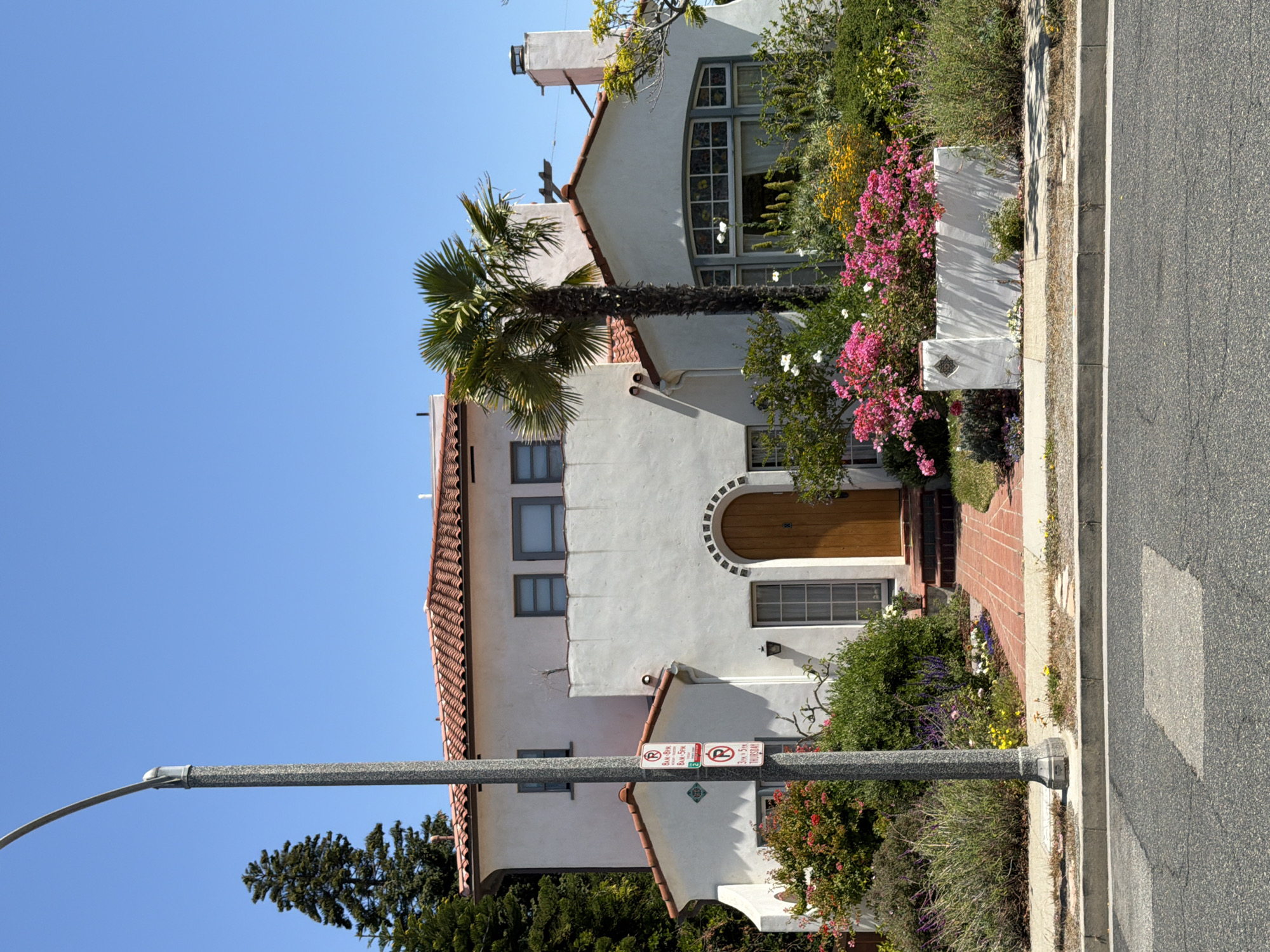 The image depicts a two-story, white stucco house with a red tile roof, characteristic of Mediterranean Revival architecture, situated in Santa Monica, California. The house features arched doorways and windows, including a distinctive arched window with stained glass on the lower level. Lush landscaping surrounds the property, with a variety of flowering plants, shrubs, and a palm tree adding to the vibrant aesthetic. A brick pathway leads to the front entrance. A street lamp stands prominently in the foreground on the left side of the image, and a sidewalk and street are visible in the lower portion of the frame. The sky is clear and blue, suggesting a sunny day. No people are visible in the image. The location is identified as Santa Monica, United States of America.