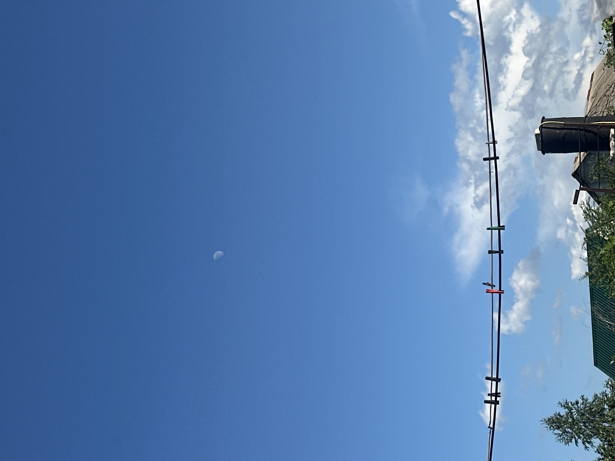 This is a vertically oriented, outdoor, daytime, medium shot photograph looking up towards the sky. The scene depicts a bright blue sky with scattered white clouds on the right side of the frame. A portion of the moon is visible as a small, pale crescent in the left half of the sky.
In the right portion of the image, a clothesline stretches diagonally upwards. Attached to the clothesline are several colorful clothespins: green, red, and dark colored ones. Behind the clothesline, a dark, cylindrical water tank is mounted on a structure, partially obscured by green foliage and the roof of a building. The building appears to have a corrugated metal roof.
The time of day appears to be daytime, likely late morning or early afternoon, given the bright sunlight and the visibility of the moon in the blue sky. The weather seems to be clear and pleasant, with only a few scattered clouds. The location context is Tobyl, Kazakhstan. No text is visible in the image.