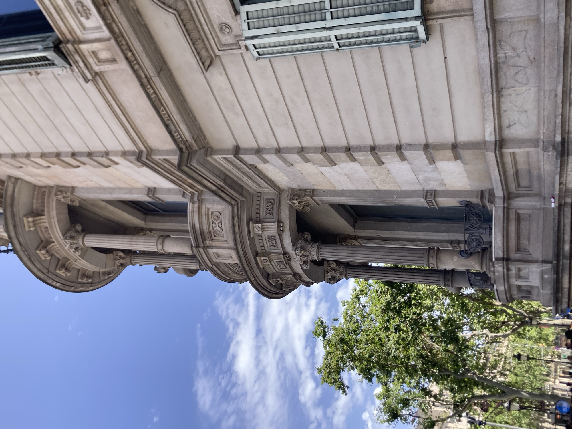 The media file captures a low-angle view of the ornate facade of a building in Barcelona, Spain. The building features classical architectural elements, including fluted columns, intricate carvings, and decorative moldings. A portion of a window with louvers is visible at the top of the frame. Below the architectural details, the bright blue sky with scattered white clouds suggests a pleasant day. A lush green tree with a dense canopy is positioned to the right of the frame, partially obscuring the view of the street below. The street level reveals glimpses of lampposts and people, indicating an urban setting with pedestrian activity. The lighting suggests it is daytime, likely mid-morning or afternoon, given the clear sky and the shadows cast by the architectural features. The overall impression is one of a historic and well-maintained urban environment. No visible text is present in the image.