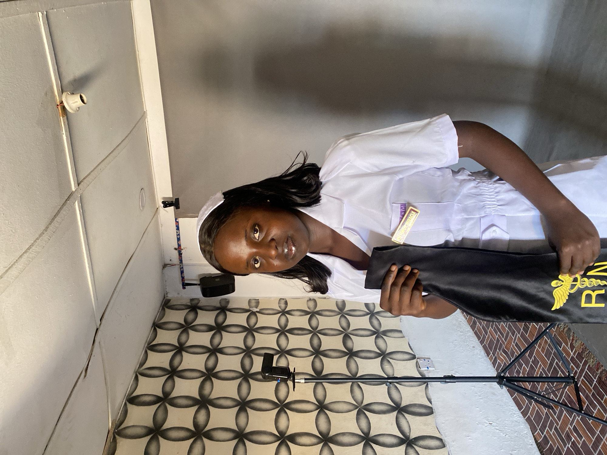 A young woman in a white nursing uniform and cap is holding a black gown with "RN" and a medical symbol embroidered in gold. She is indoors. The floor is tiled with a geometric pattern. A lighting rig is visible. A bare lightbulb socket is attached to a wall. The location is Vom, Nigeria.