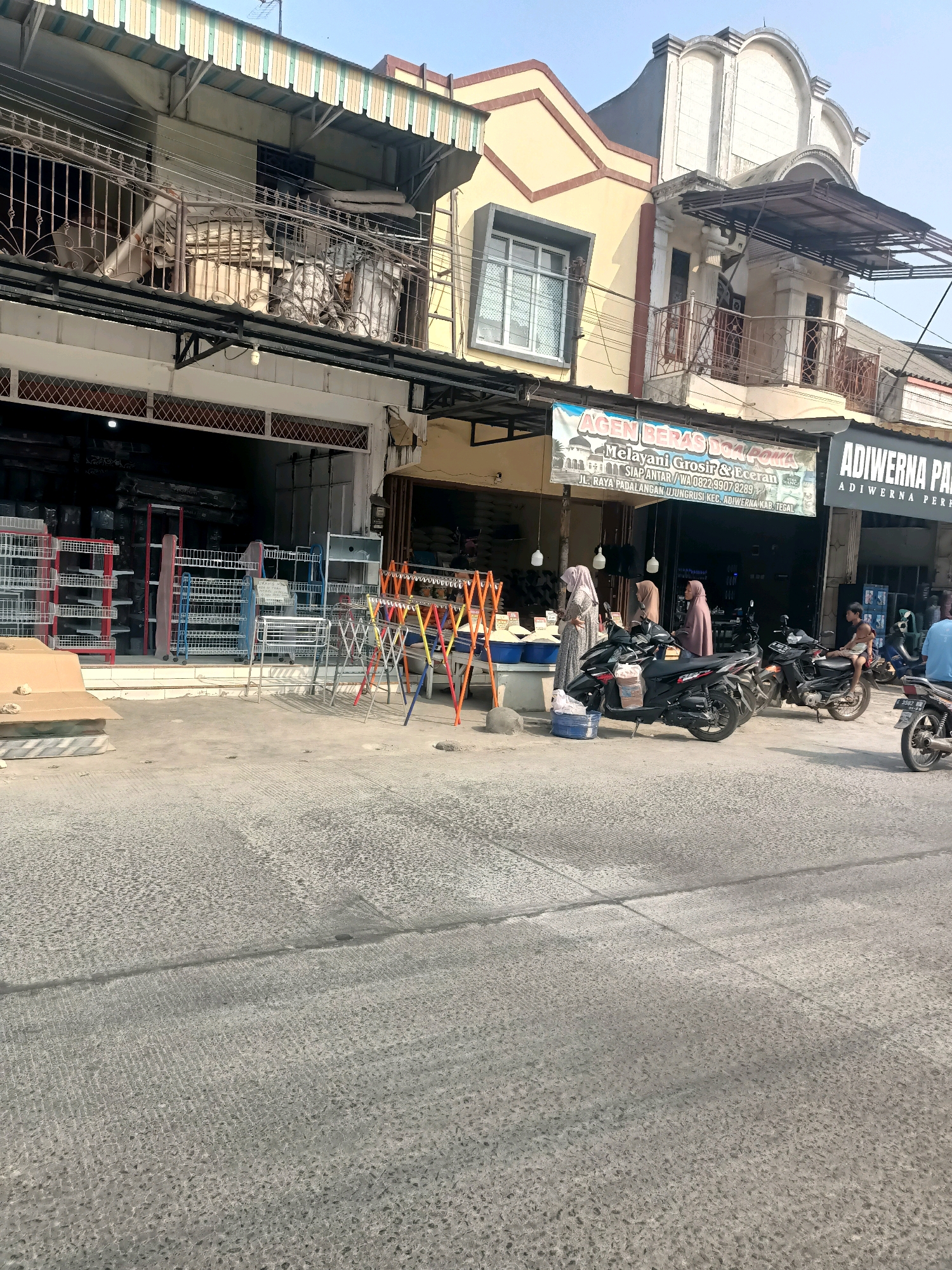 The image depicts a street scene in the city of Tembok Banjaran, Indonesia, with a focus on the storefronts and the street in front of them. On the left, there is a building with several levels, featuring balconies with railings and awnings. Below the balconies are storefronts with metal racks displaying various items, possibly household goods or store fixtures. A large cardboard box sits on the ground in front of one of the storefronts.
To the right, a banner hangs above a storefront that reads "AGEN BERAS DOA POEMA Melayani Grosir & Eceran" and further text in Indonesian. This indicates a rice agency that serves both wholesale and retail customers. The storefront itself is open, revealing shelves stocked with items.
In front of the buildings, several motorcycles are parked. Three women, dressed in hijabs and long clothing, are visible near the "AGEN BERAS DOA POEMA" storefront, with one woman appearing to interact with another or perhaps a display. One motorcycle is black and is parked in front of the storefront, while another is black and silver is positioned further to the right.
A young boy is sitting on the back of a motorcycle on the right side of the image, and a person in a blue shirt is walking away from the camera on the far right. The street in the foreground is paved with textured concrete, showing signs of wear and repair. The overall environment suggests a commercial area in a moderately populated urban setting. The sky is clear and blue.