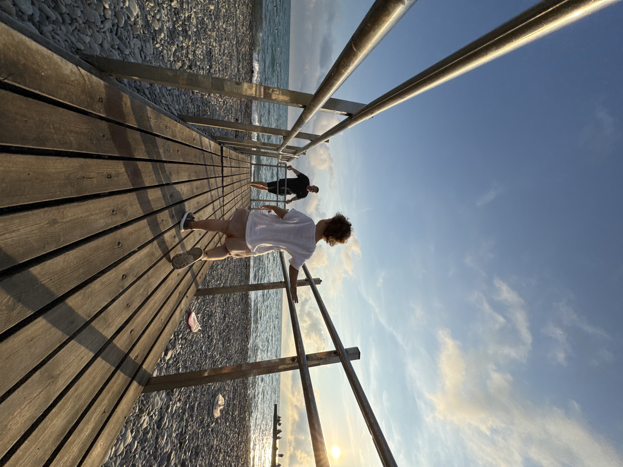 A low-angle, wide-shot perspective captures a wooden boardwalk extending over a pebble beach toward the sea, bathed in the warm glow of late afternoon sunlight. Two figures are present on the boardwalk. In the foreground, a child, facing away from the camera and towards the sea, walks forward. They wear a white t-shirt and light-colored shorts. Further down the boardwalk, a man stands with his hands on a metal railing, facing the child. He is dressed in a black t-shirt and dark shorts. The sea is visible to the left, with gentle waves lapping the shore. The sky above is a clear blue with scattered, wispy clouds. In the distance, at the end of the boardwalk, a group of silhouetted figures can be seen near the water's edge. The scene is likely on a beach in Sochi, Russia, given the architecture of the boardwalk and railing, common in resort towns along the Black Sea coast.