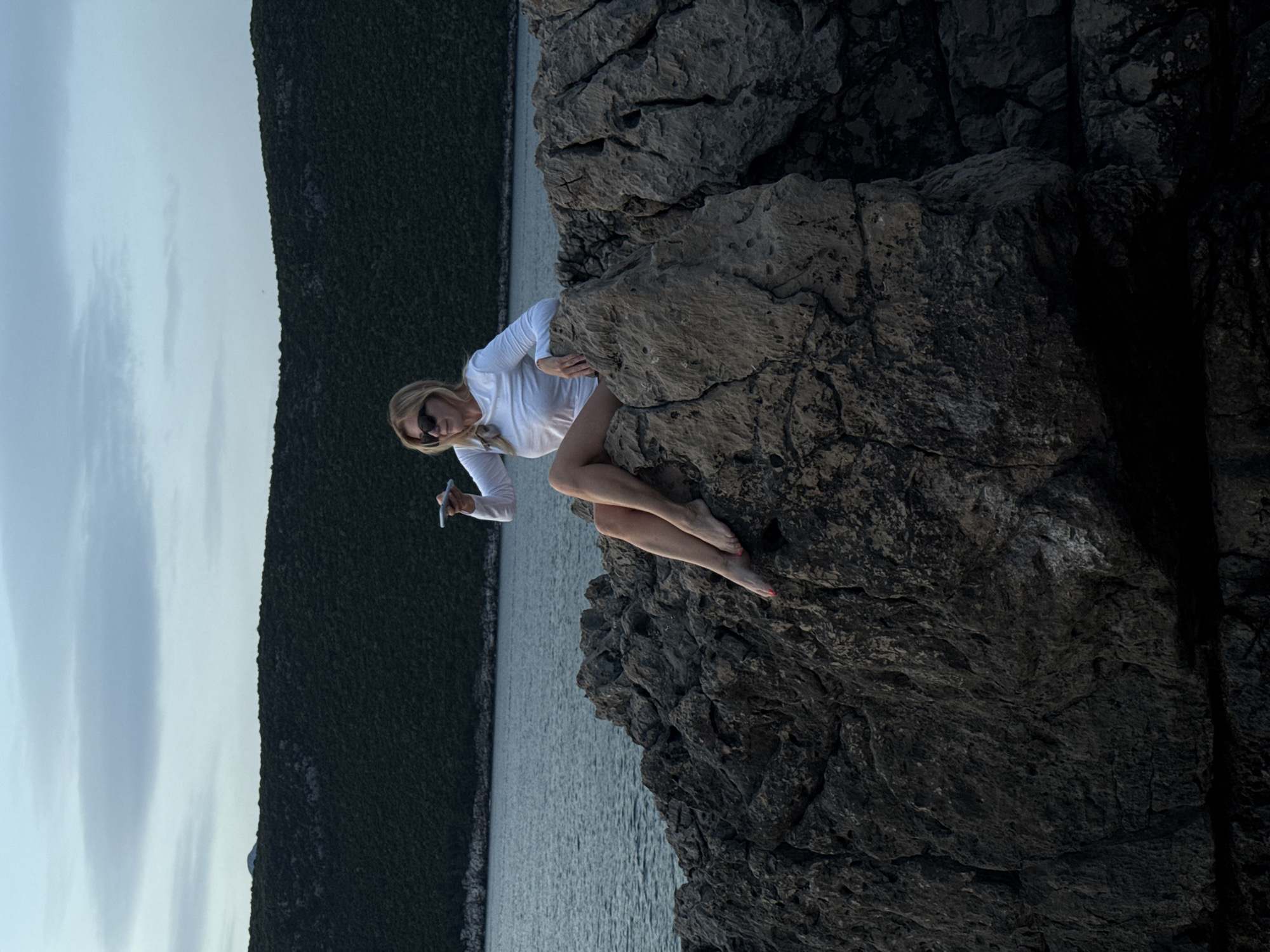 The image captures a woman with blonde hair and sunglasses, dressed in a white long-sleeved shirt, sitting barefoot on dark, textured rocks. She appears to be holding a mobile phone. The background consists of a calm body of water with a distant, tree-covered shoreline, transitioning into a cloudy sky. The overall scene suggests an outdoor setting in Luštica, Montenegro, with the woman positioned on coastal rocks.