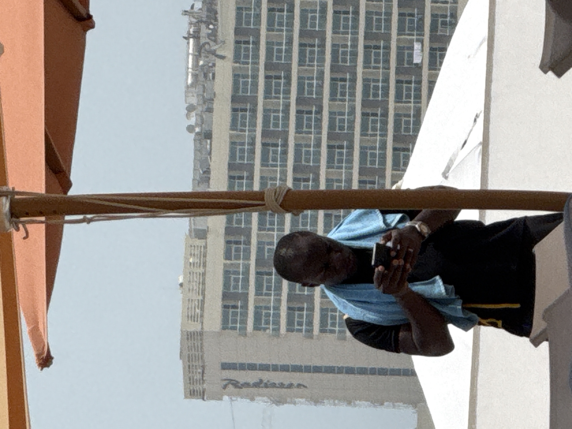 A dark-skinned man stands outdoors, looking down at his smartphone. He wears a black t-shirt and a light blue towel is draped around his neck and shoulders. A gold-toned wristwatch is visible on his left wrist. He is positioned in front of a large, modern building with many windows, and behind him, further in the distance, is another taller building with similar window patterns. A portion of an orange umbrella and a wooden pole are in the foreground, suggesting the man is under shade. The sky appears hazy and bright, indicating daylight. The environment suggests a warm climate, possibly a resort or urban area with tall structures.