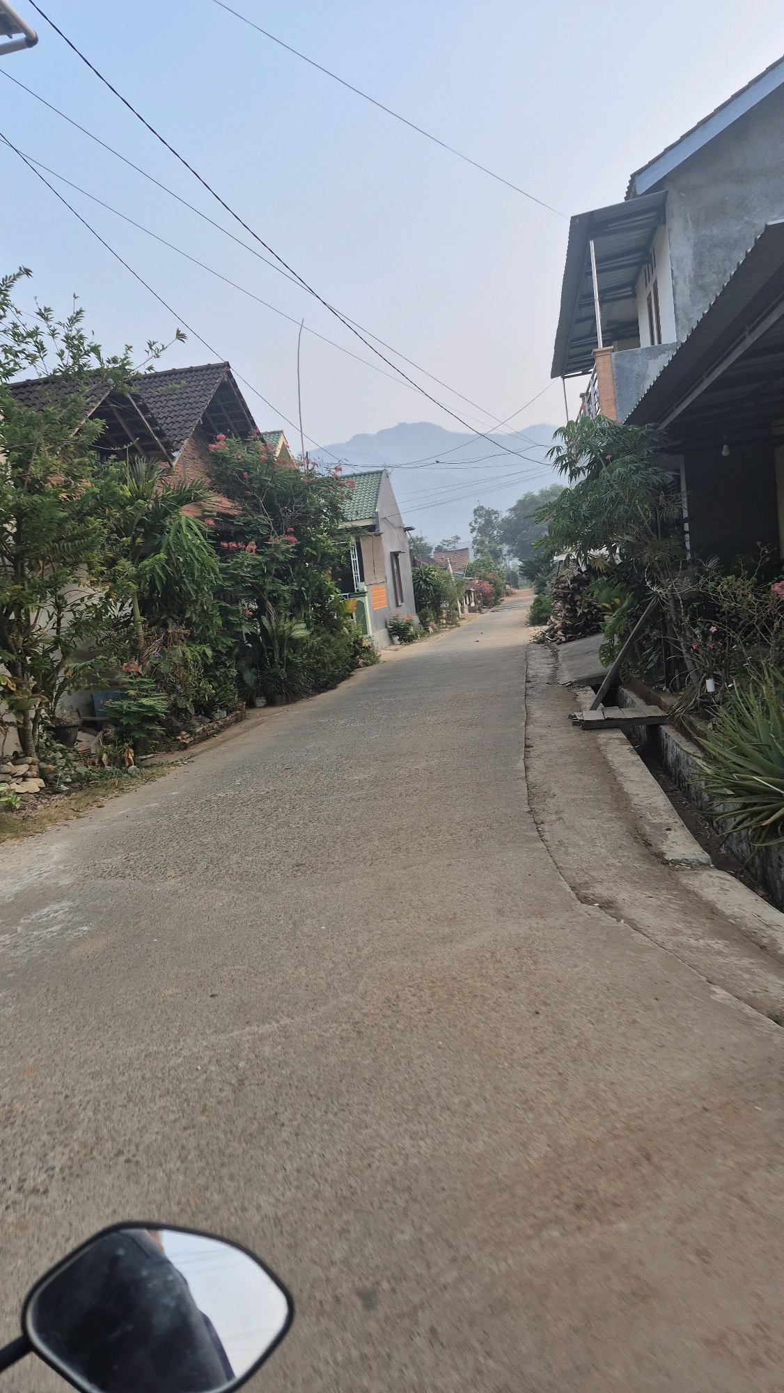 The image shows a view from a motorcycle as it travels down a paved road in a rural or suburban area, likely in Wiroko, Indonesia, given the architectural style of the houses and the lush vegetation. The road curves gently to the right and recedes into the distance, bordered by houses on both sides and dense greenery. The houses have pitched roofs, some with dark tiles and others with green or lighter colored roofs. Many of the houses are adorned with abundant plants, including flowering bushes and large leafy plants, suggesting a tropical or subtropical climate. Overhead, a network of power lines crisscrosses the sky. In the background, distant hills are visible, shrouded in a light mist or haze, indicating early morning or late afternoon light conditions. The road surface is textured asphalt. In the foreground, the reflection of the rider and the motorcycle's side mirror are visible in the lower left corner. The overall atmosphere is tranquil and suggests a peaceful, everyday scene in a residential area.