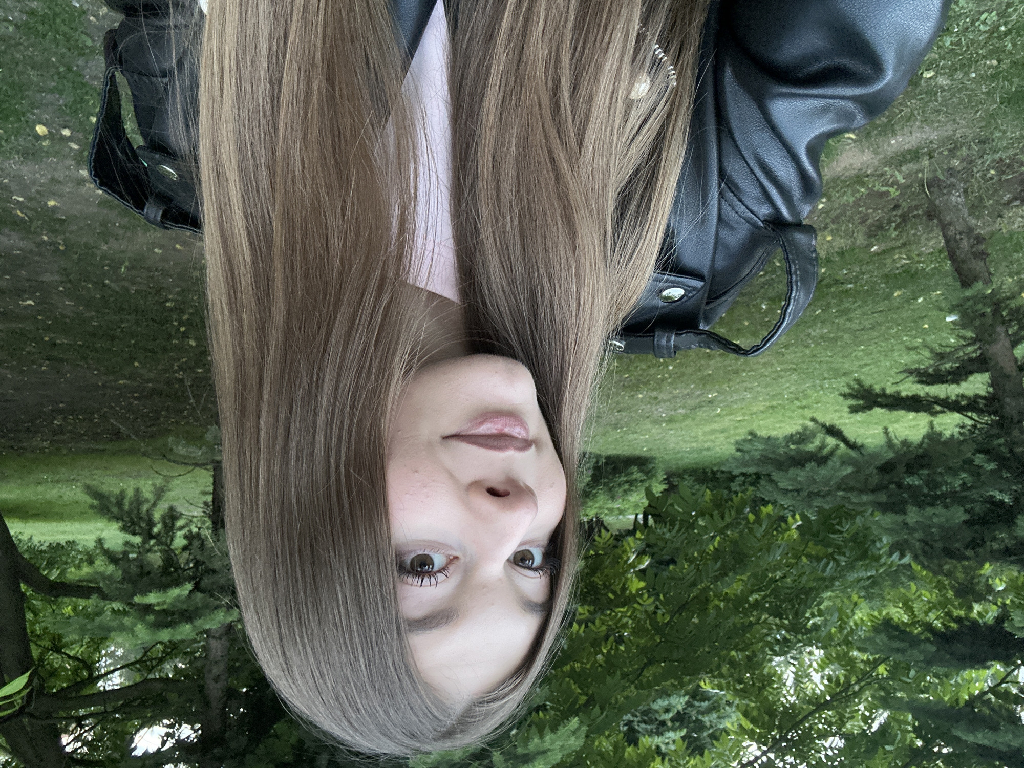 This close-up self-portrait, originally oriented upside down, features a young woman looking directly at the viewer. Her long, straight, light brown hair frames her face, extending past her shoulders. She has brown eyes and a neutral to slightly contemplative expression. She is wearing a black leather jacket, identifiable by its texture, silver hardware (snaps, buckles), and a collar, over a light pink top visible beneath her hair.
The setting is outdoors, in what appears to be a park or garden in Kazan, Russia. The background is filled with lush green foliage, including bright green grass and a variety of trees with both dark and lighter green leaves and needles. The lighting suggests it is daytime, with soft, even illumination and no harsh shadows, indicating either an overcast day or a period of soft light like late afternoon. No specific activity beyond posing for a photo is depicted. There is no visible text in the image.