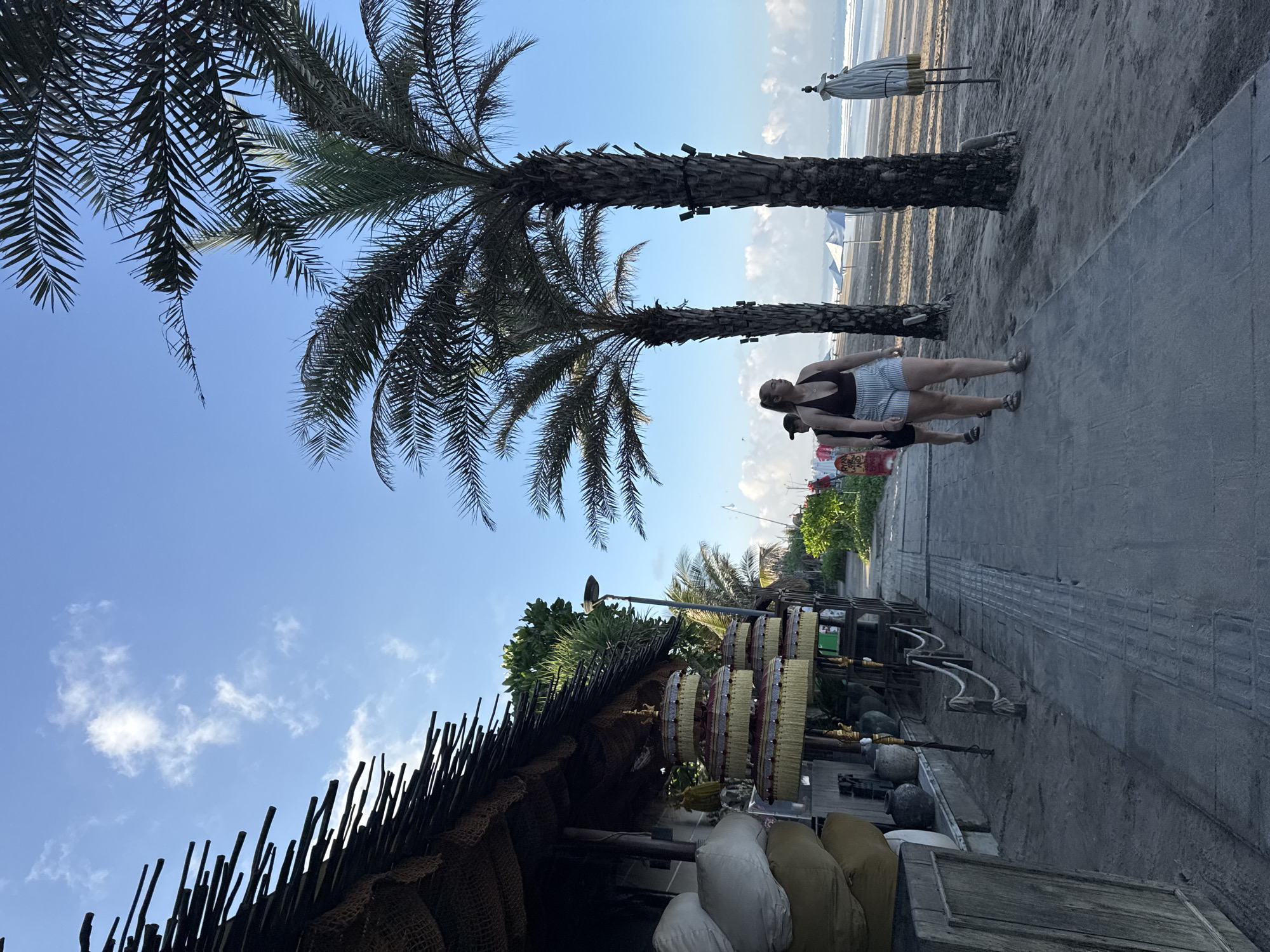 This media file depicts a tropical beach scene in Kuta, Indonesia, characterized by a paved pathway, lush palm trees, and a rustic stall, with the ocean and sandy beach visible in the background.
Two individuals are captured walking along the pathway. In the foreground, a woman with dark hair wears a dark sleeveless top, striped shorts, and sandals, looking towards her left. Partially obscured behind her is another person wearing a dark cap and shirt. They appear to be casually strolling.
To the left, a roadside stall features a roof constructed from woven materials and sticks. It displays several stacks of what appear to be colorful, round objects, possibly fruits or traditional offerings. Below the display, sacks or cushions are arranged alongside ceramic pots. The pathway is made of grey pavers. Tall palm trees with broad fronds line the path, their trunks showing textures and signs of age.
Beyond the pathway and trees, the sandy beach stretches towards the horizon, where gentle waves can be seen. A folded beach umbrella is present on the sand, with other beach structures faintly visible in the distance. The sky is clear and blue with scattered white clouds, indicating a bright daytime setting, possibly late morning or early afternoon due to the soft, even lighting. Text is present on a small sign near the stall but is too blurry to be legible.