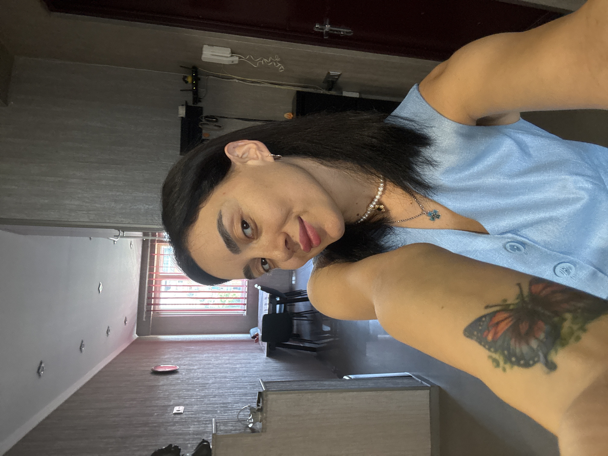 The image is a close-up, selfie-style portrait of a woman with dark hair, wearing a light blue top and a pearl necklace. She is looking towards the camera with a slight smile. A butterfly tattoo is visible on her right arm. The background appears to be an indoor setting, possibly a room in an apartment or house, with a window showing a glimpse of trees and a building. The room has grey walls and carpeted floors, with some furniture visible, including chairs and a table. The lighting suggests it could be daytime. The overall mood is casual and relaxed. The image is oriented sideways, suggesting it was taken with a phone.