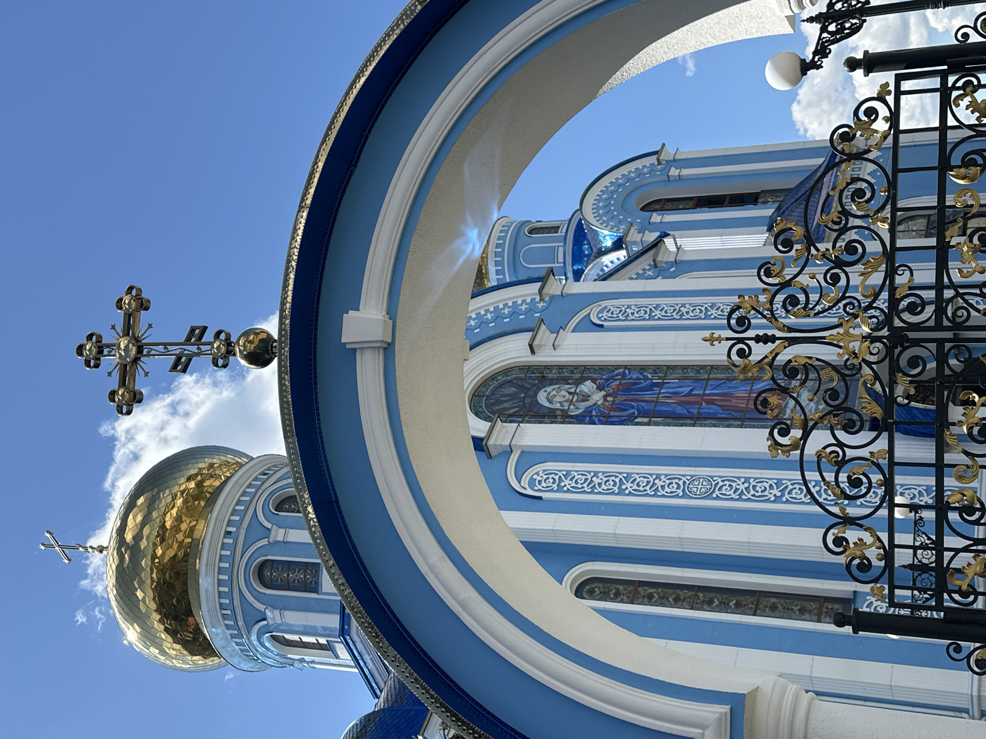 This is a vertical, eye-level, full shot of the Church of the Resurrection in Zhovte, Ukraine, on a clear day. The church is white and blue with a golden dome and a tall cross on top. A large blue and white archway frames the entrance to the church. In front of the church is a black metal gate with gold embellishments. The sky is blue with a few white clouds. The image is focused on the church and its architectural details.