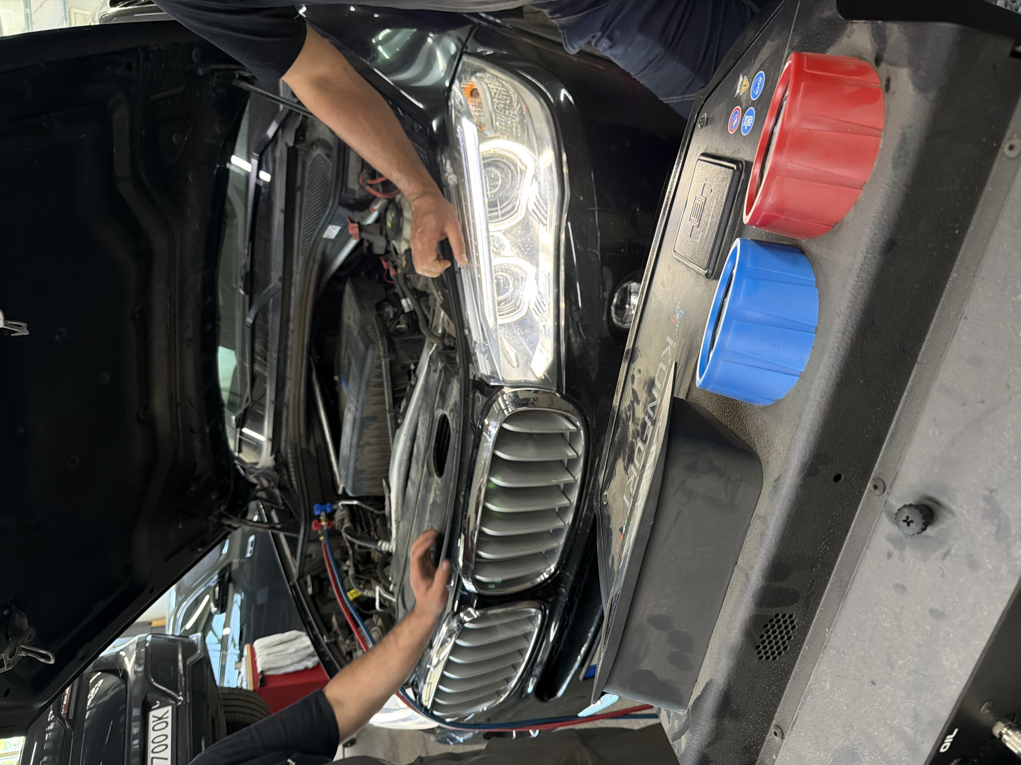 In a dimly lit garage in Burgas, Bulgaria, a black BMW SUV with its hood open is undergoing maintenance. The engine is partially exposed, with various hoses and components visible. Two individuals, likely mechanics, are attending to the vehicle. One person's arm is seen reaching towards the engine bay, while another person's arms are positioned near the front of the car, possibly working on the lights or grill. In the foreground, a diagnostic machine or tool cart is present, with two colored caps (one blue, one red) resting on its surface. The machine is dusty and shows signs of wear. The overall scene suggests a routine car repair or diagnostic procedure taking place.