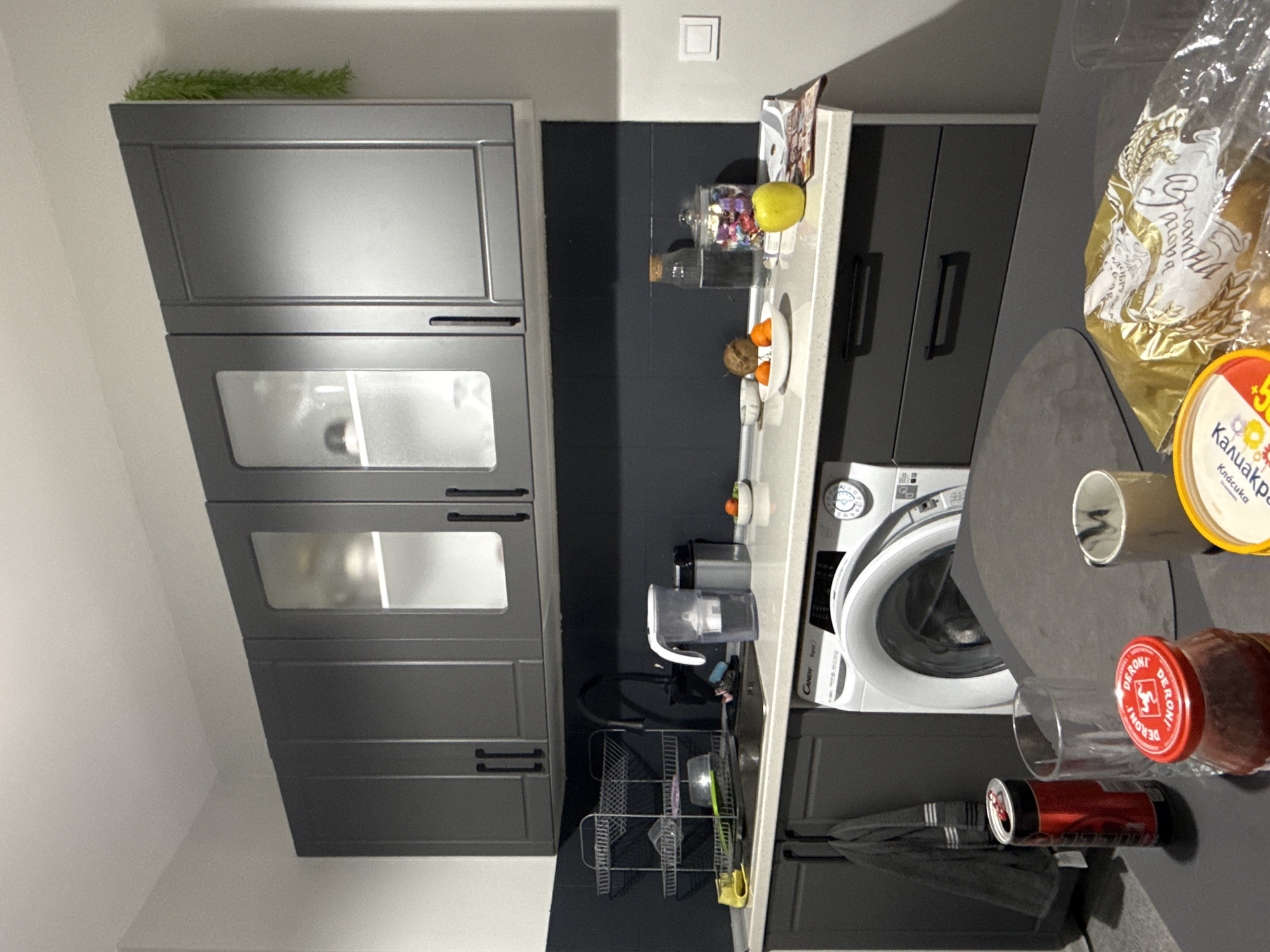 A domestic interior is presented from an elevated perspective, showcasing a kitchen setup. The scene is oriented with the kitchen cabinets on the left, progressing towards the right where various objects are arranged on a countertop. The color scheme includes gray cabinets with glass-paneled doors, a speckled white countertop, and black tiled walls. A washing machine is situated under the countertop. Food items, utensils, and cleaning supplies are scattered on the countertop. No people are visible. The location in the city of Stara Zagora, Bulgaria is not discernible.