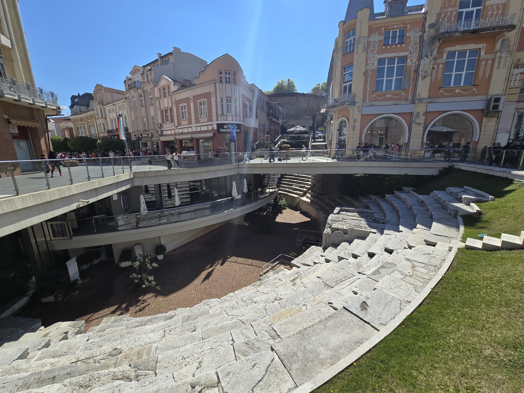 This image shows an ancient amphitheater in Plovdiv, Bulgaria, integrated within the modern urban landscape. The amphitheater consists of rows of stone seating descending toward a central performance area. Above the amphitheater is a modern pedestrian bridge lined with glass railings, allowing people to walk over the site. Buildings with varying architectural styles stand on either side of the bridge. Some people are visible on the bridge, observing the amphitheater and its surroundings. Below the bridge is a modern structure with glass windows and outdoor seating, likely a restaurant or cafe. The overall environment is a mix of historical ruins and contemporary architecture.