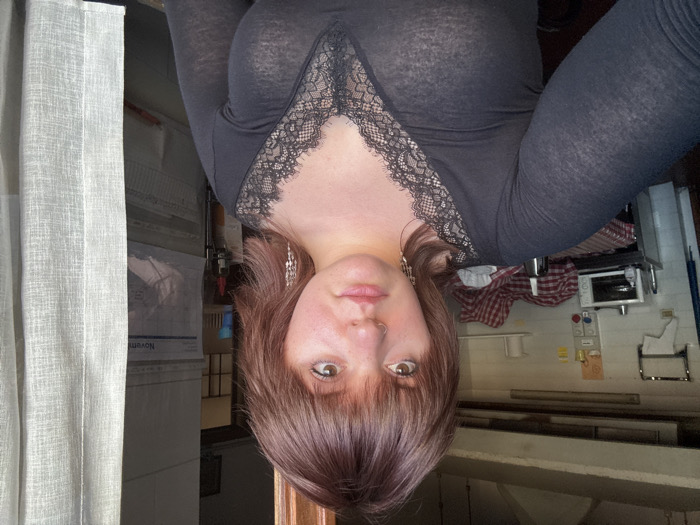 A female individual, appearing to be in her 20s or early 30s with reddish-brown, shoulder-length hair and bangs, is centered in an indoor setting in Cortona, Italy. She wears a dark, sheer, long-sleeved top with a black lace V-neckline and long, multi-element dangle earrings. A small stud nose ring is visible on her left nostril, and her expression is neutral as she looks directly forward. To her left, the background shows a functional kitchen or utility area with light-colored walls, a metal towel rack, a counter covered by a red and white checkered tablecloth, a small microwave or toaster oven, a bottle, and crumpled red and white patterned fabric. To her right, a doorway or opening is partially obscured by a light-colored, sheer curtain. A wall calendar displaying "Novem" (November) is visible through the opening. Natural light illuminates the scene, seemingly from the right side of the frame.