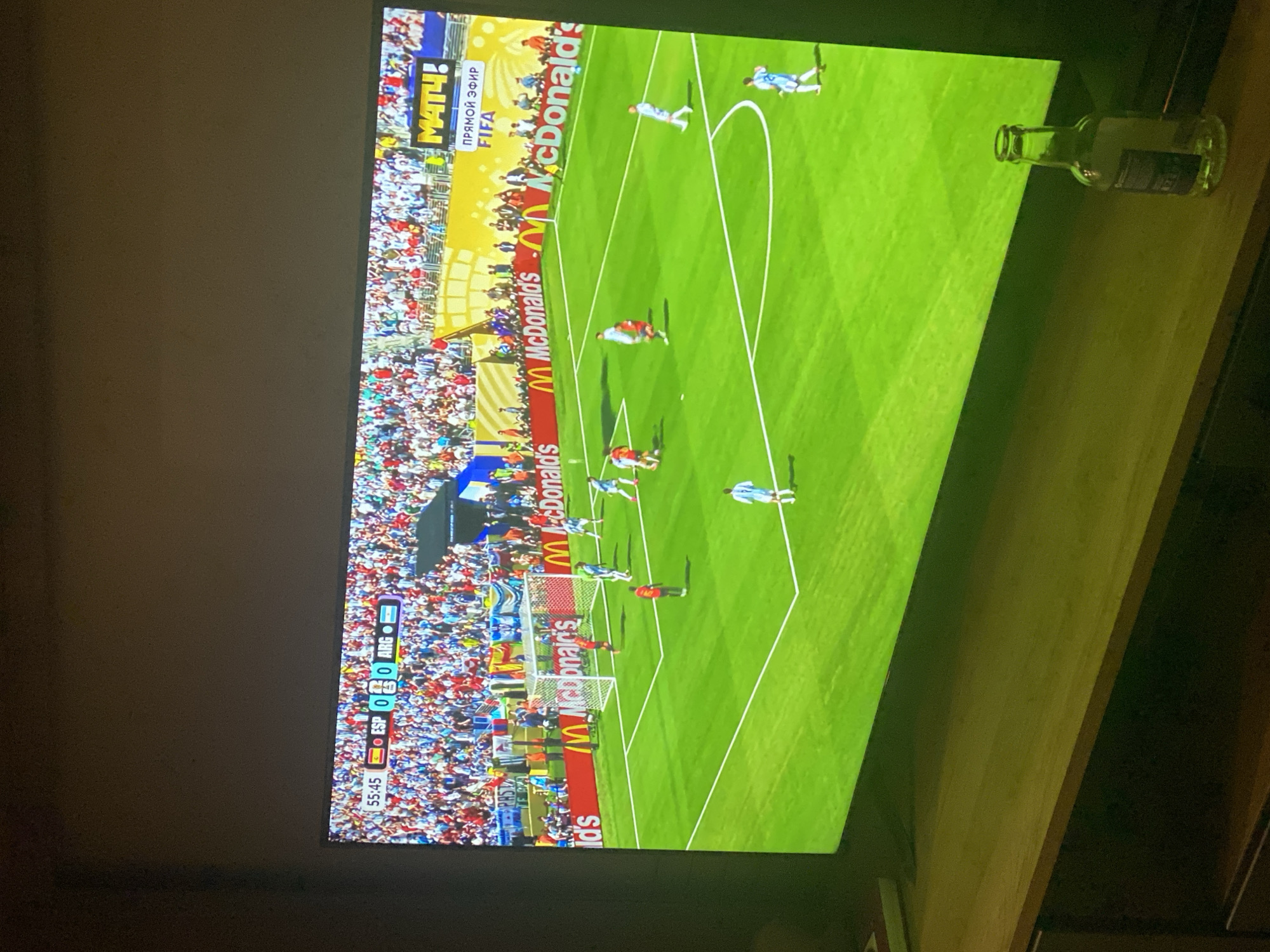 The image shows a television screen displaying a soccer match between Spain (ESP) and Argentina (ARG) with the score at 0-0 in the 55th minute and 45 seconds. The game appears to be a simulation or part of a video game, judging by the graphics and the "MATH!" and "ПРЯМОЙ ЭФИР FIFA" (Live Broadcast FIFA) overlays on the screen, suggesting it is being broadcast in Moscow, Russia. The field is bright green with white markings. Several players in light blue and red uniforms are visible on the field, with some near the goalposts. A large crowd of spectators, wearing a variety of colorful clothing, is visible in the stadium stands behind the field. In the foreground, on a wooden surface in front of the television, sits a clear glass bottle. The overall lighting suggests an indoor environment, possibly a living room or entertainment area, with the television being the primary light source.
