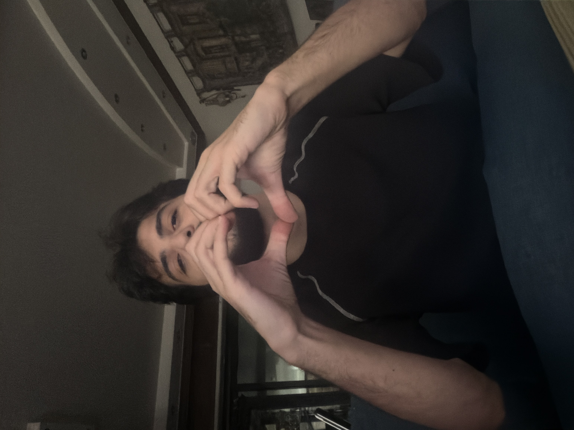 A medium-shot, slightly overhead, waist-up image shows a young man with dark hair and a beard, wearing a black t-shirt, forming a heart shape with his hands in front of his face. The man is seated on a dark blue surface, likely a couch, indoors. The background is dimly lit, with a dark ceiling featuring recessed lighting and a decorative cornice. To the right, a framed artwork hangs on a wall. The environment appears to be a residential setting, possibly in the Sarouja Municipality, Syria, given the hints. The image quality is somewhat grainy.