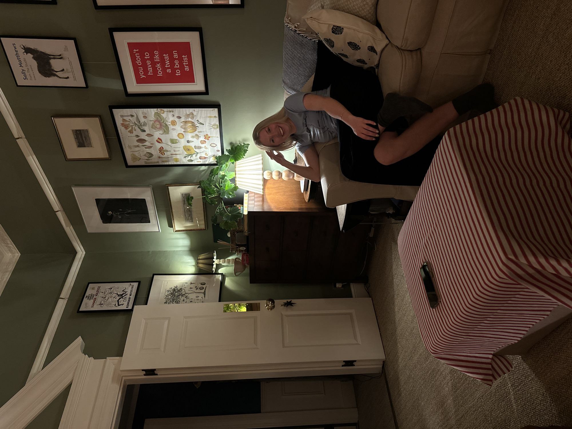 Here's a detailed description of the image:
**Scene:** Interior of a living space, possibly a house or apartment. The setting is warm and dimly lit, suggesting evening or low light conditions.
**People:** A young woman with blonde hair is seated on a beige couch, appearing relaxed and engaging with the viewer, perhaps posing for the picture. She is wearing casual clothing, including what appears to be dark leggings and a light-colored t-shirt.
**Objects:**
* A couch is prominent in the scene. It is covered with a striped red and white blanket.
* A dark wood chest of drawers is placed against the wall.
* A lamp is placed on the chest of drawers.
* Numerous framed pictures and posters are hung on the wall behind the couch and chest of drawers. The posters include the text "Sally Matthews", "you don't have to look like a twat to be an artist". "SUMMER SOLSTICE, BLOOMSBURY WAITER'S RACE"
* A doorway is visible in the lower part of the frame, leading into another room that appears to be dark.
* On the top of the striped cover, there is what appears to be a phone
**Environment:** The walls are painted a muted, dark green. The overall decor suggests a bohemian or artistic taste.
**Location:** Based on the information contained within the posters, the location is London, United Kingdom.