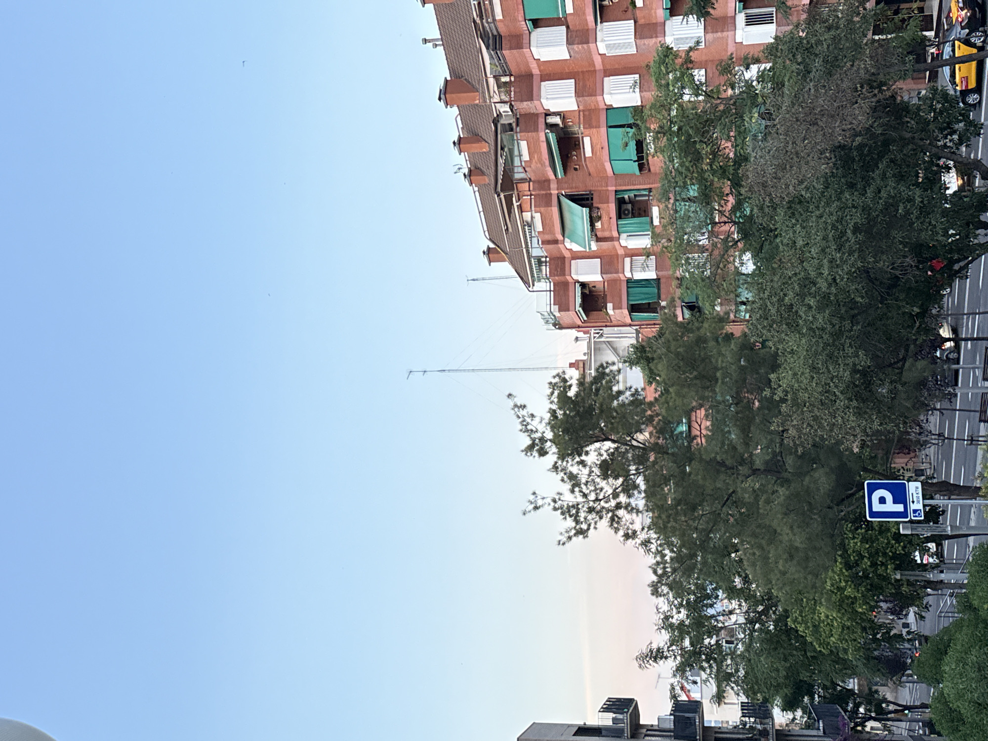 The image depicts a street scene in Barcelona, Spain, featuring a residential building and surrounding trees. The building, with its red brick facade and numerous windows adorned with green awnings and white shutters, dominates the right side of the frame. The sky above is a clear, pale blue, with hints of pink and orange near the horizon, suggesting either sunrise or sunset.
In the foreground and middle ground, lush green trees of various sizes create a canopy over the street. A blue parking sign with a white "P" and an accessibility symbol is visible on the left side of the street. A yellow taxi with black accents is parked or moving along the road, indicating urban activity. The street itself is paved, and there are glimpses of other vehicles and buildings in the background, hinting at a densely populated urban environment.
The overall impression is of a typical city street in Barcelona, bathed in soft, natural light.