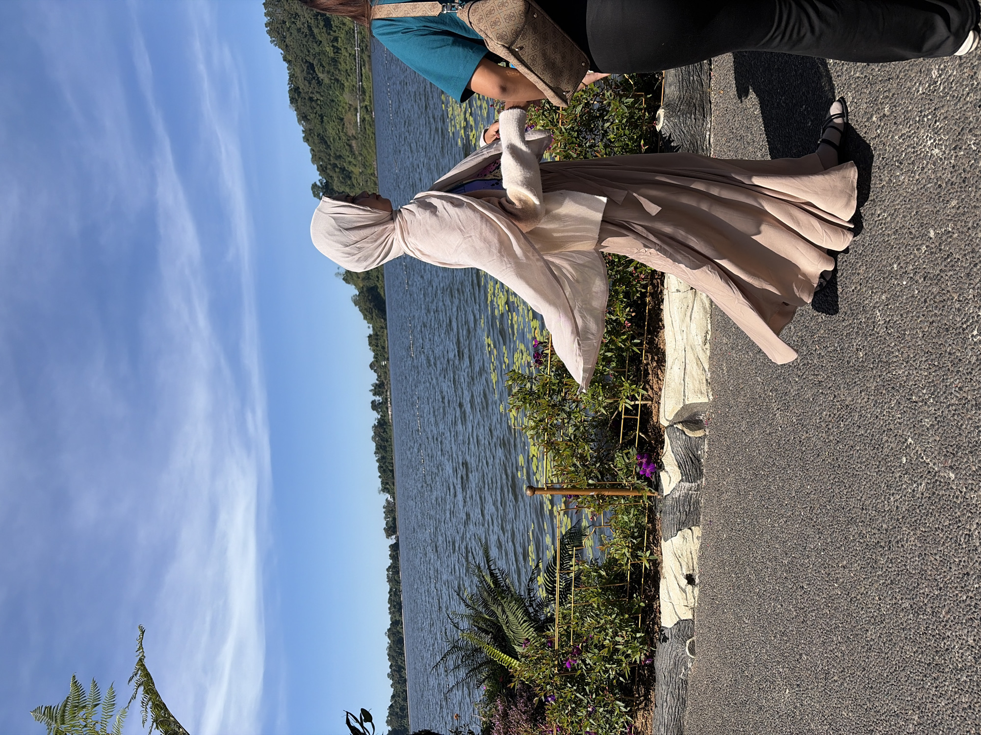 This is a full-shot, eye-level, outdoor photograph of two women walking on a paved path near a lake in Baturiti, Indonesia. The woman on the left is wearing a beige hijab and a long beige dress that is flowing in the wind. She is walking away from the camera, looking to her left. The woman on the right is partially visible from the waist down, wearing a teal shirt and black pants, and carrying a brown patterned crossbody bag. Her head and face are not visible. The background features a large body of water with some lily pads, a lush green hill covered in trees, and a bright blue sky with wispy white clouds. In the foreground, there is a decorative border made of grey and white stones. The overall scene depicts a serene and sunny day by the lake.
