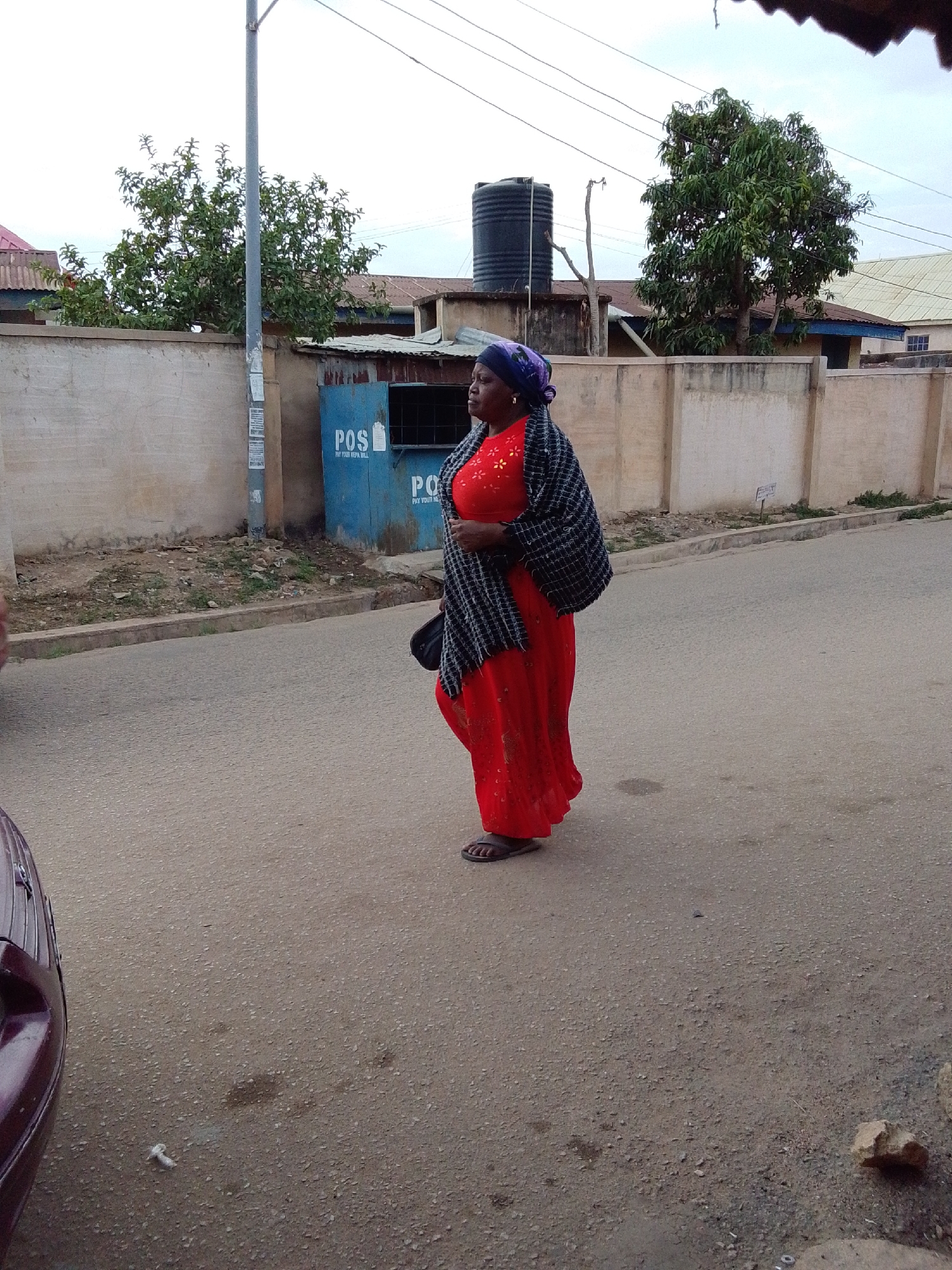The image captures a street scene featuring a woman walking on a paved road. The woman, positioned slightly right of center, wears a long, loose-fitting red dress adorned with small, scattered yellow or orange patterns. A dark, possibly black and white, checkered or striped shawl is draped over her shoulders, and she has a dark purple head covering. She is wearing dark sandals and appears to be holding a black bag or purse with her left hand, which is not fully visible.
In the background, a light-colored concrete wall separates the road from residential or commercial buildings. These structures have varied roofs, including corrugated metal. Two green trees with dense foliage are visible above the wall, one on the left and a larger one on the right. A black water tank stands on a platform behind the wall, near the right tree. A tall, light-grey utility pole is prominent on the left side of the image, bearing some faded white notices.
To the right of the utility pole and behind the woman, two small, blue-painted kiosks or booths are visible. The left kiosk prominently displays the text "POS" (Point of Sale), with smaller text beneath that clearly reads "PAY YOUR NEPA BILL". The second kiosk to its right also shows partially visible text, starting with "PO".
The road surface is asphalt with a scattering of small gravel. To the far left in the foreground, the maroon or dark red fender of a car is partially visible. The area between the road and the wall is a dirt shoulder with some sparse dry vegetation and debris. The sky above is overcast, suggesting daytime with subdued light.