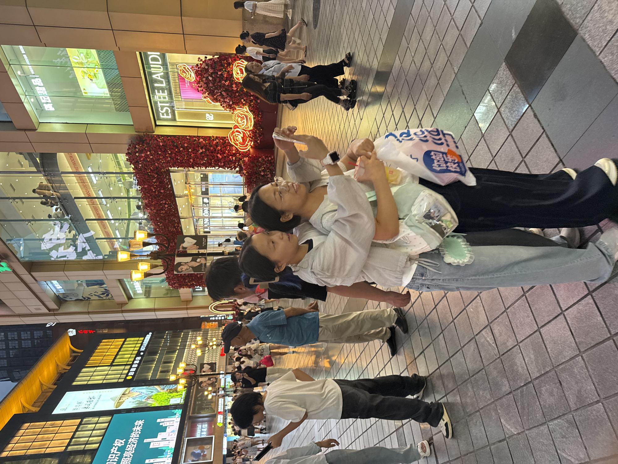 A street scene in the Yuzhong District of China at dusk shows a group of people walking on a tiled pedestrian walkway. The buildings on either side of the street are brightly lit with neon signs and displays. In the foreground, two young women are interacting, with one taking a picture of the other with a phone. They are holding shopping bags. Other pedestrians are visible in the background, including men and women of various ages. Some are walking alone, while others are in groups. The street is clean and well-maintained, and the overall atmosphere appears to be lively and bustling. The architecture of the buildings suggests a modern urban environment, with large glass windows and multi-story structures. Decorative floral arrangements are prominent on some of the buildings, adding a touch of color to the scene. The lighting indicates the transition from daylight to evening, with streetlights and building lights casting reflections on the wet pavement.