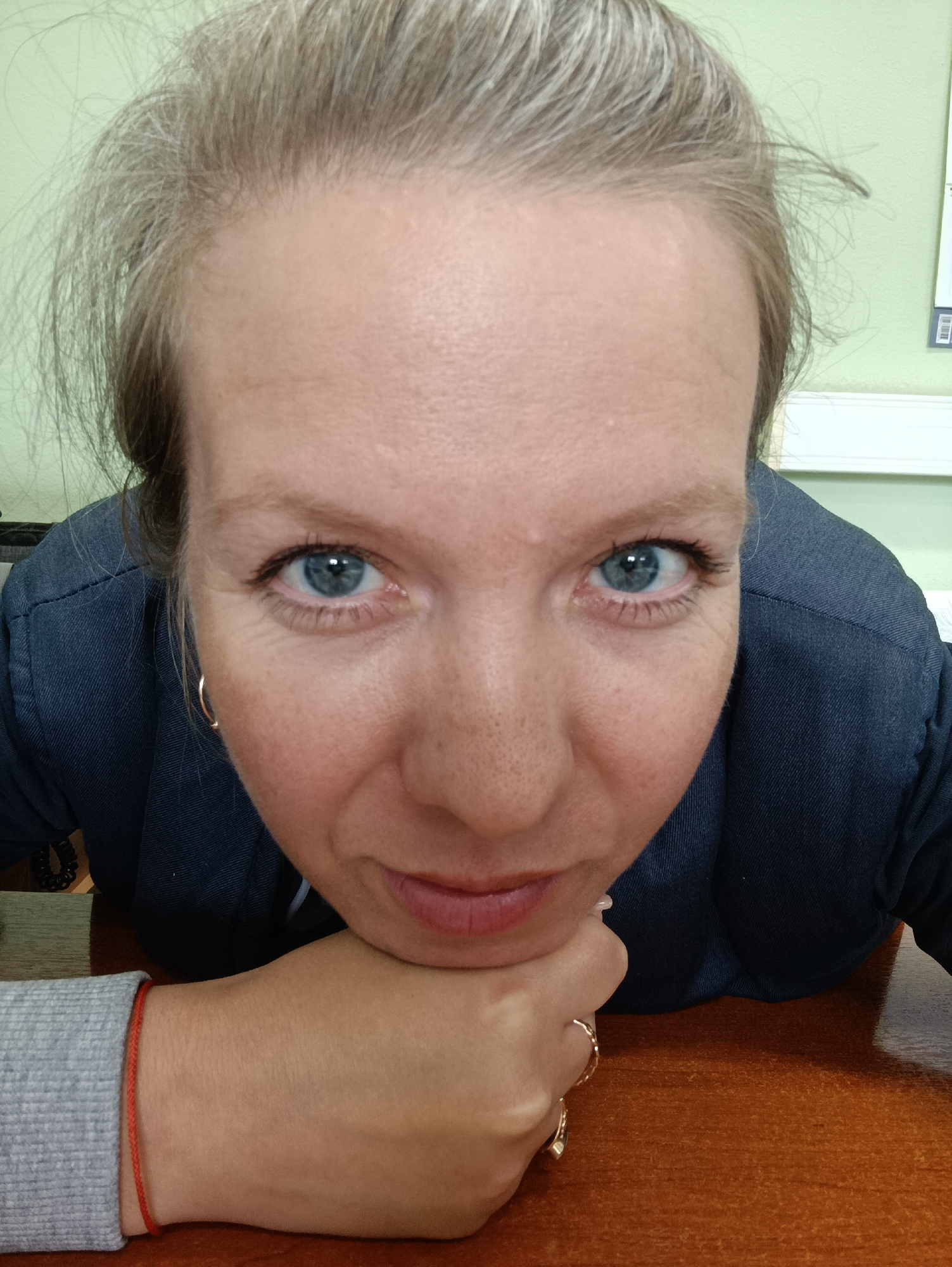 A close-up, high-angle photograph depicts a woman resting her chin on her hand. She has light blonde hair, fair skin, and light blue eyes. She is wearing a dark blue blazer over a grey long-sleeved shirt or sweater. She has a red bracelet on her left wrist and a gold ring on her left hand. The background includes a light green wall. The photo was taken on a brown wooden surface, likely a table. The exact location within the city of Chaikovsky, Russia is indeterminable from the photograph.