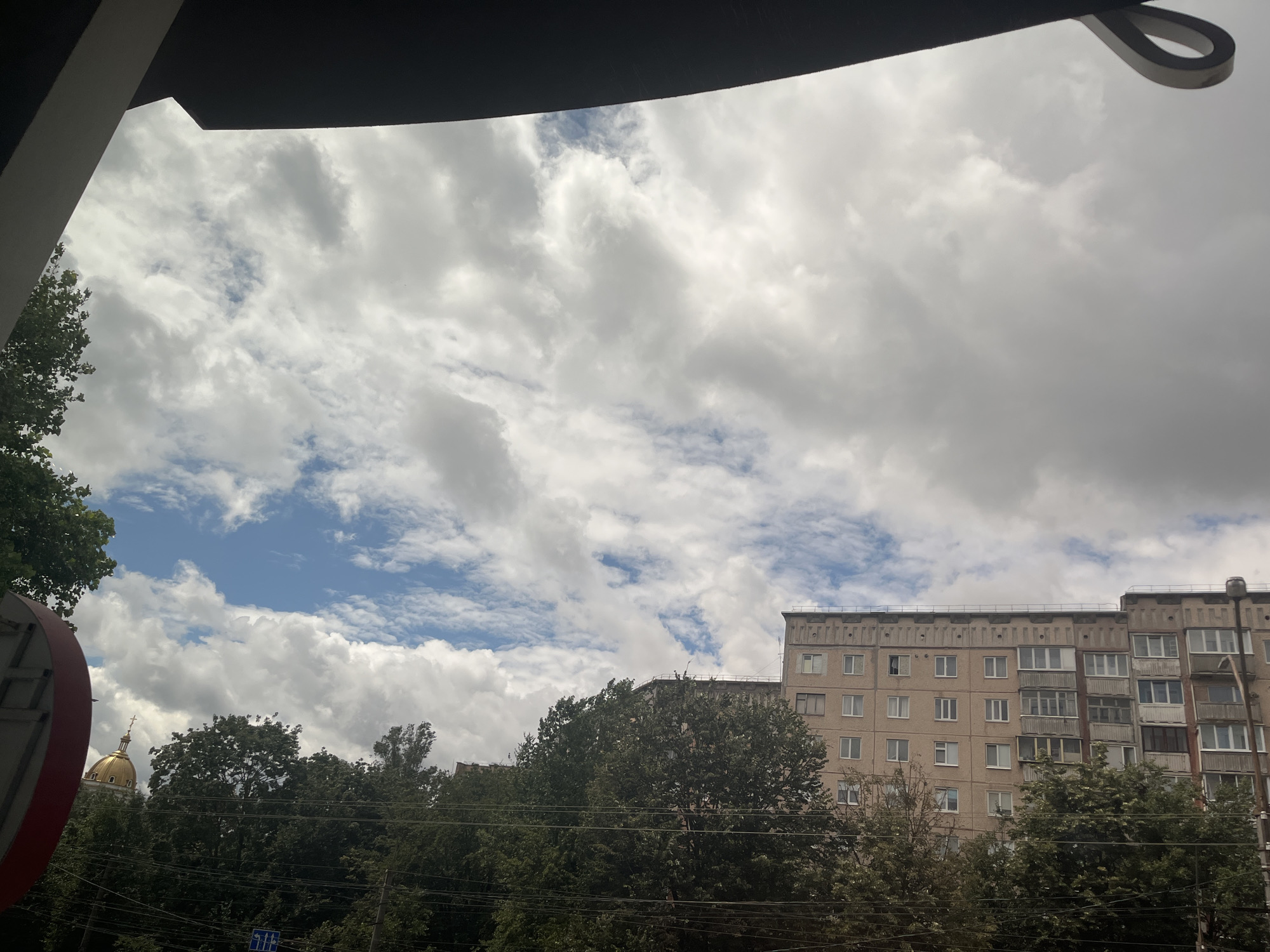 A daytime, eye-level exterior shot captures a view of the sky above a residential building and trees. The sky is filled with an abundance of white, fluffy clouds interspersed with patches of blue. In the middle ground, a multi-story, pale beige apartment building with numerous windows is visible, its architecture suggesting a Soviet-era design. Below the building, a dense canopy of dark green trees forms a thick line across the scene. To the left, a glimpse of a golden dome with a cross on top can be seen peeking through the trees, hinting at a church or cathedral nearby. The foreground is partially obscured by a large, dark, curved overhang, possibly part of a building or a structure. Power lines are visible running horizontally across the lower portion of the image.