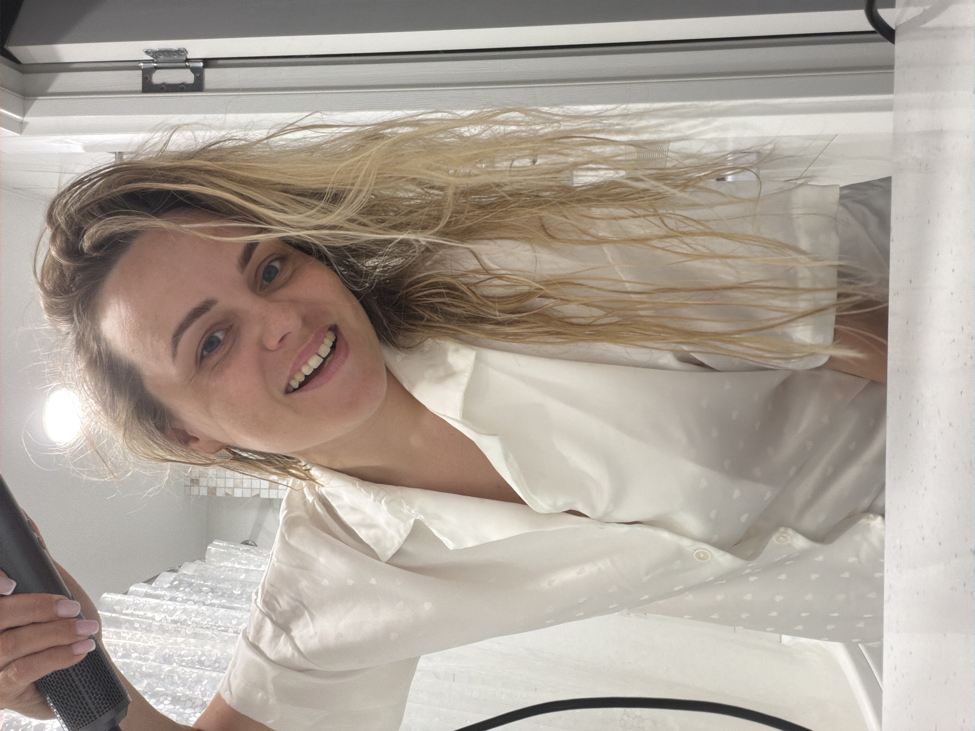 A woman with blonde hair is holding a microphone and smiling at the camera. She is wearing a white shirt with small dots. The background includes a white wall with square tiles, a ceiling, and a window. The location is not identifiable as Votkinsk, Russia.