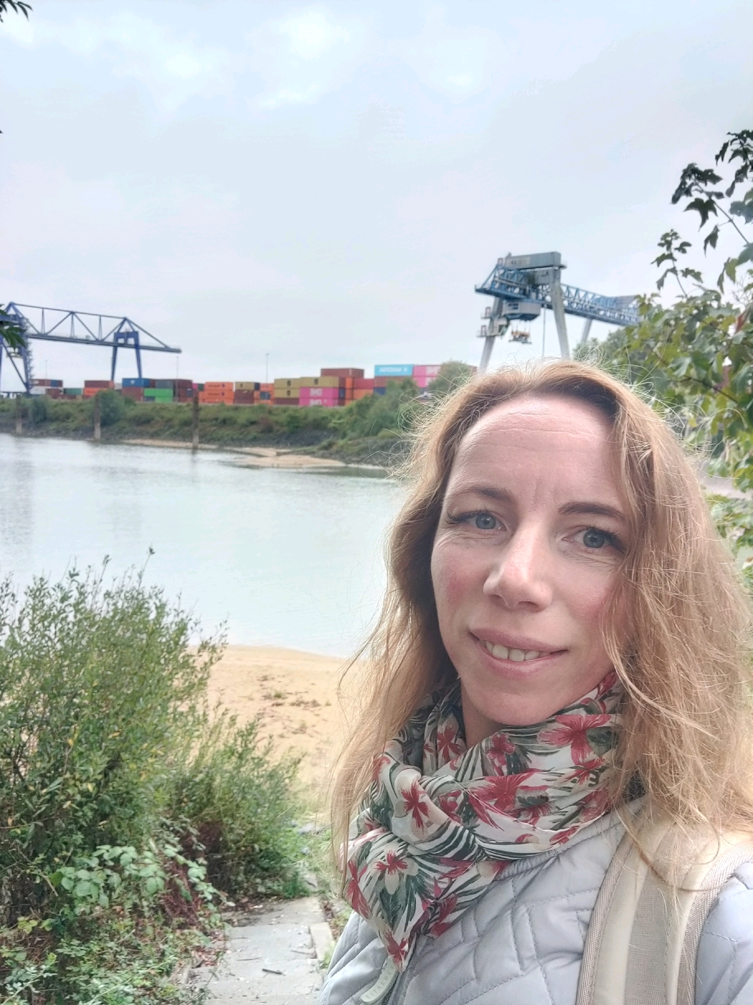 An outdoor, medium shot selfie features a blonde woman with blue eyes and fair skin, smiling at the camera. She is wearing a light grey quilted jacket and a floral scarf with red and white flowers. Her hair is shoulder-length and appears windswept.
The background depicts a waterfront industrial scene. A wide, calm body of water is in the mid-ground, with a sandy bank visible on the left. Across the water, a green embankment is lined with colorful shipping containers stacked high. Two large industrial cranes, one blue and one grey, are visible in the distance, suggesting a port or shipping facility. The sky is overcast with grey clouds. Green foliage and trees are present on the right and left sides of the frame, framing the central scene. The overall atmosphere is somewhat muted due to the cloudy weather, but the woman's smile adds a touch of warmth to the image. The scene is likely located in Emmerich on the Rhine, Germany, given the presence of the Rhine river and industrial port activities.