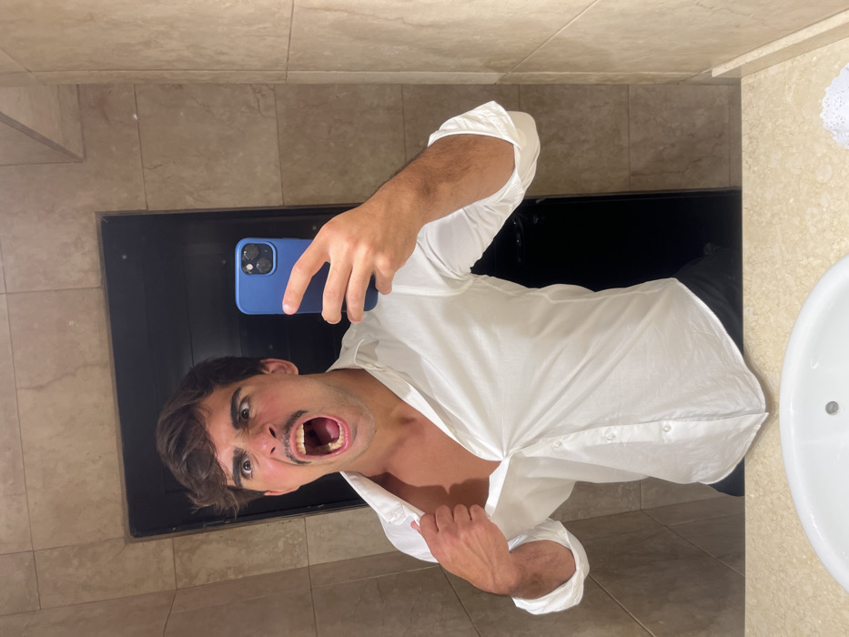 A man in a white shirt is taking a selfie in a bathroom. He has his mouth open wide in a comical expression. He is holding a blue phone up to the mirror. The bathroom has tan tiles and a black framed mirror. A white sink is visible in the reflection.