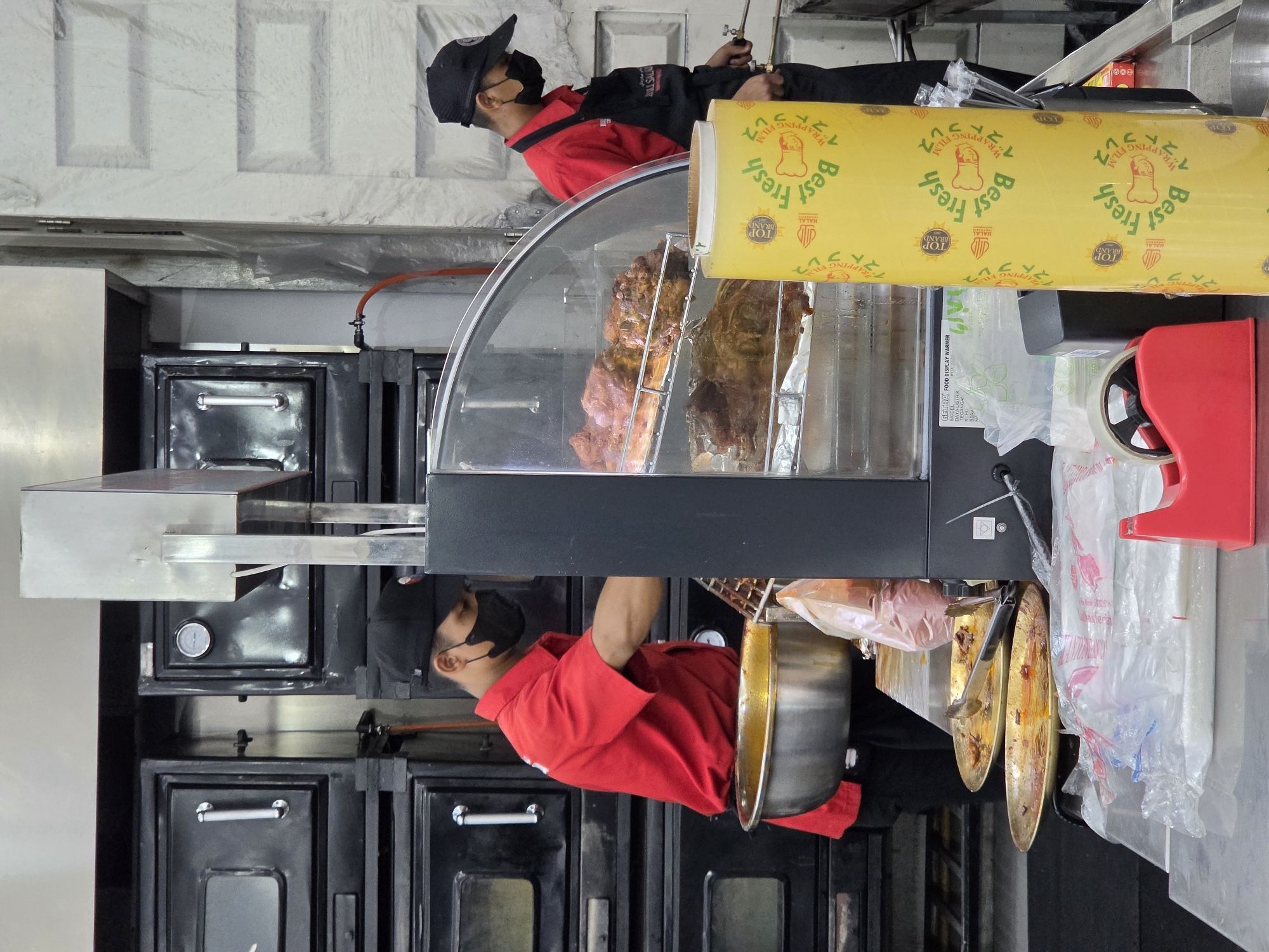 This image captures a scene within a food establishment, likely a restaurant or food stall in Medan, Indonesia. The primary subjects are two individuals, both wearing face masks and red uniforms, suggesting they are employees. One individual, in the foreground, is wearing a black cap and a red shirt. They are leaning over a large, cylindrical metal pot, possibly preparing or serving food. A second individual, in the background and also wearing a black cap and red shirt, appears to be interacting with a large roll of yellow plastic wrap.
The setting is a commercial kitchen or food preparation area. Visible objects include kitchen appliances such as ovens or warming units, a glass display case showcasing large cuts of meat, and various containers and utensils. The yellow plastic wrap prominently features text in both English and what appears to be Japanese characters, along with graphics indicating "Best Fresh," "TOP BRAND," and "HALAL." This suggests the establishment caters to a diverse clientele and emphasizes the freshness and religious compliance of their products.
The overall impression is one of a busy food service environment. The lighting suggests an indoor setting, likely during operating hours. The presence of the meat in the display case and the preparation activities indicate that food is actively being handled and served. There are no strong emotional cues visible due to the face masks, but the actions suggest a focus on work and customer service. The visible text on the packaging provides brand and product information, reinforcing the commercial nature of the scene.