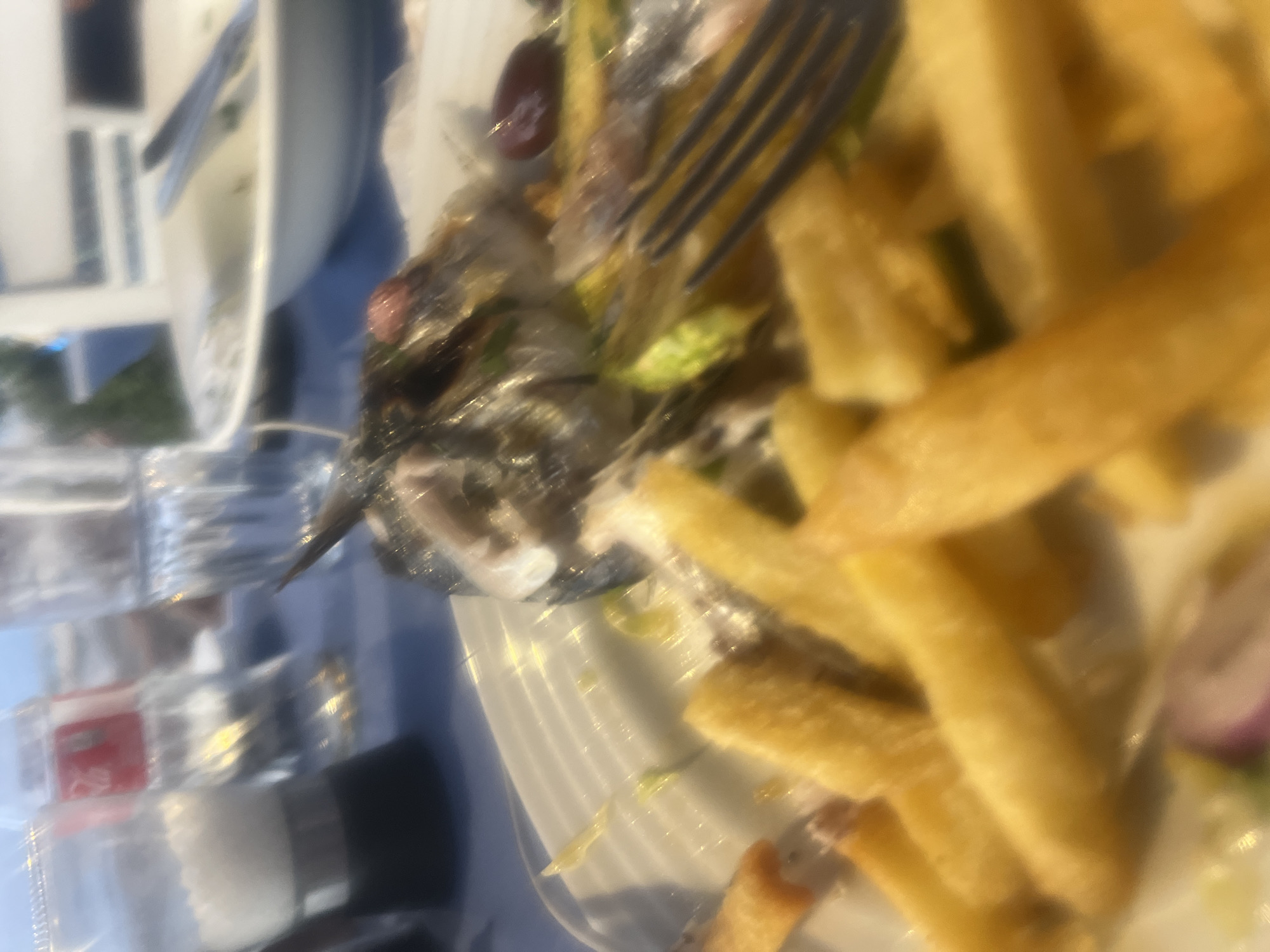 The image captures a close-up view of a partially eaten fish dish served with French fries on a white plate. The fish appears to be mostly consumed, with only the head and some bones remaining. A fork is visible, resting on the plate and partially obscuring the fish. The French fries are golden brown and scattered around the plate. In the background, out-of-focus elements suggest an outdoor setting, possibly a restaurant, with a table covered in a blue tablecloth, salt and pepper shakers, a bottle of Coca-Cola, and glassware. The scene is likely from Polis Chrysochous, Cyprus, given the context of the prompt.