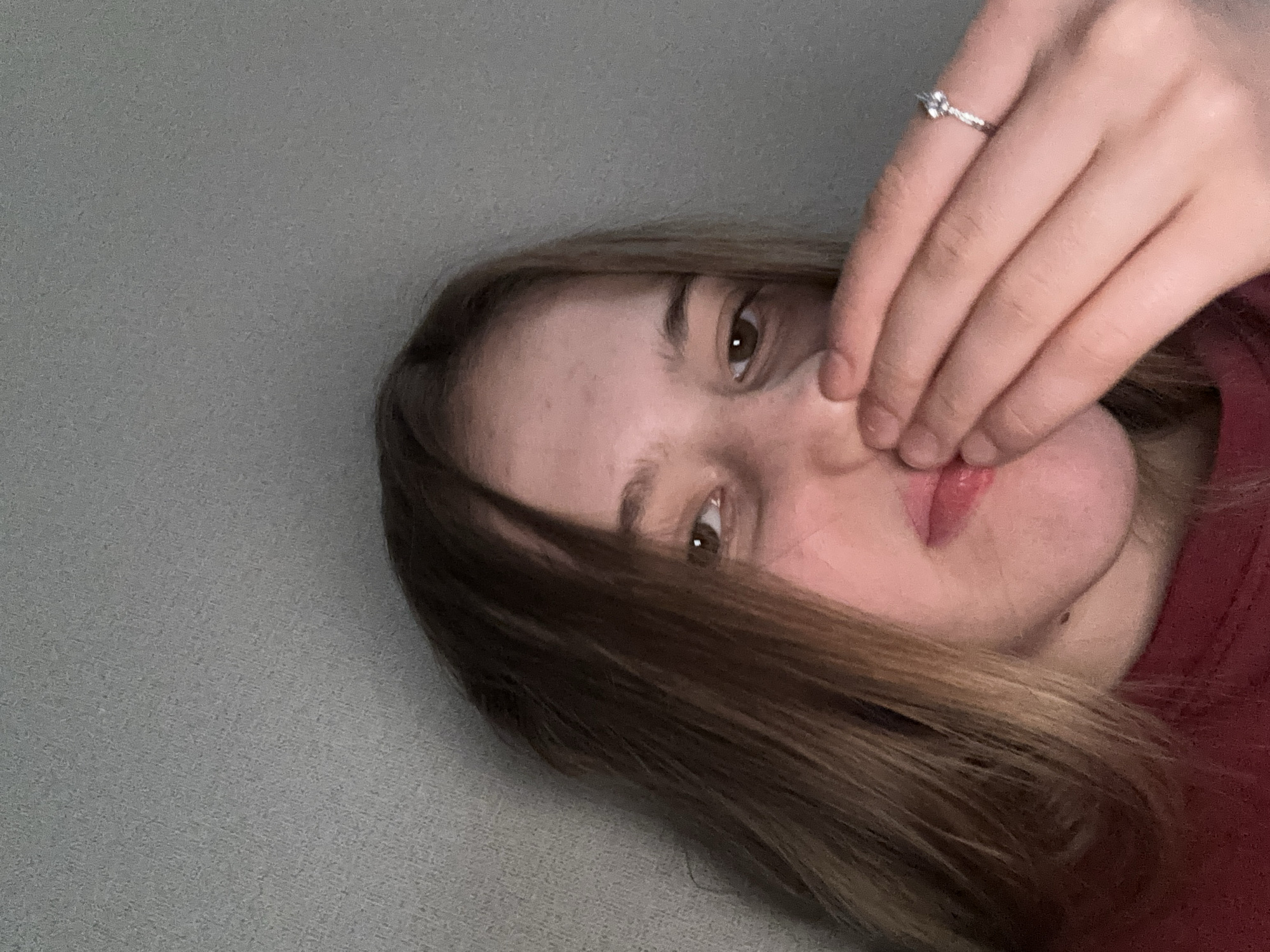 The image shows a close-up of a young woman's face, partially obscured by her hand. She has light brown hair and is wearing a dark red shirt. A ring with a small stone is visible on her right ring finger. The background is a textured gray wall. The image appears to have been taken indoors in the city of Sinyavino, Russia.