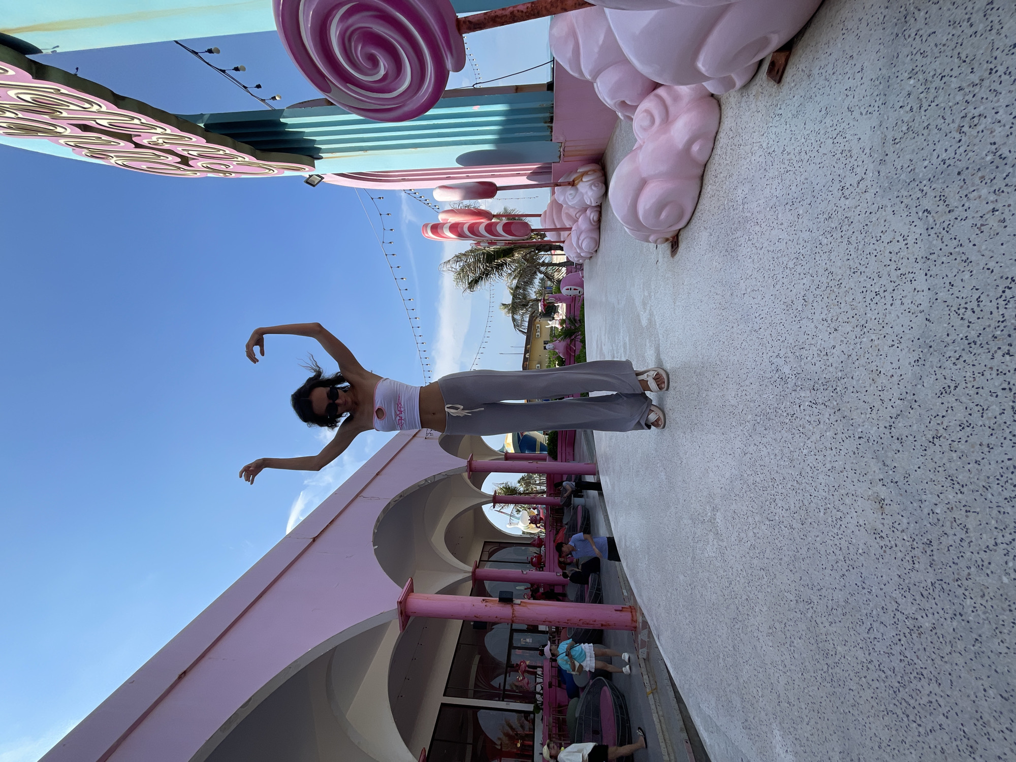 In Tien Thanh Ward, Vietnam, a young woman stands with her arms outstretched in a playful pose on a white, speckled pavement. She is wearing a white bandeau top with a pink cutout and gray wide-leg pants. Behind her, a pink building with arched openings and columns creates a vibrant backdrop. To her right, a large blue structure with a pink sign is adorned with oversized lollipop decorations in shades of pink and purple, along with pink cloud-like sculptures. The scene is illuminated by bright daylight under a clear blue sky, with string lights visible in the background. Several people are scattered in the distance, some near the pink building, others further down the walkway. Palm trees are visible behind the lollipop decorations, suggesting a tropical or coastal location.