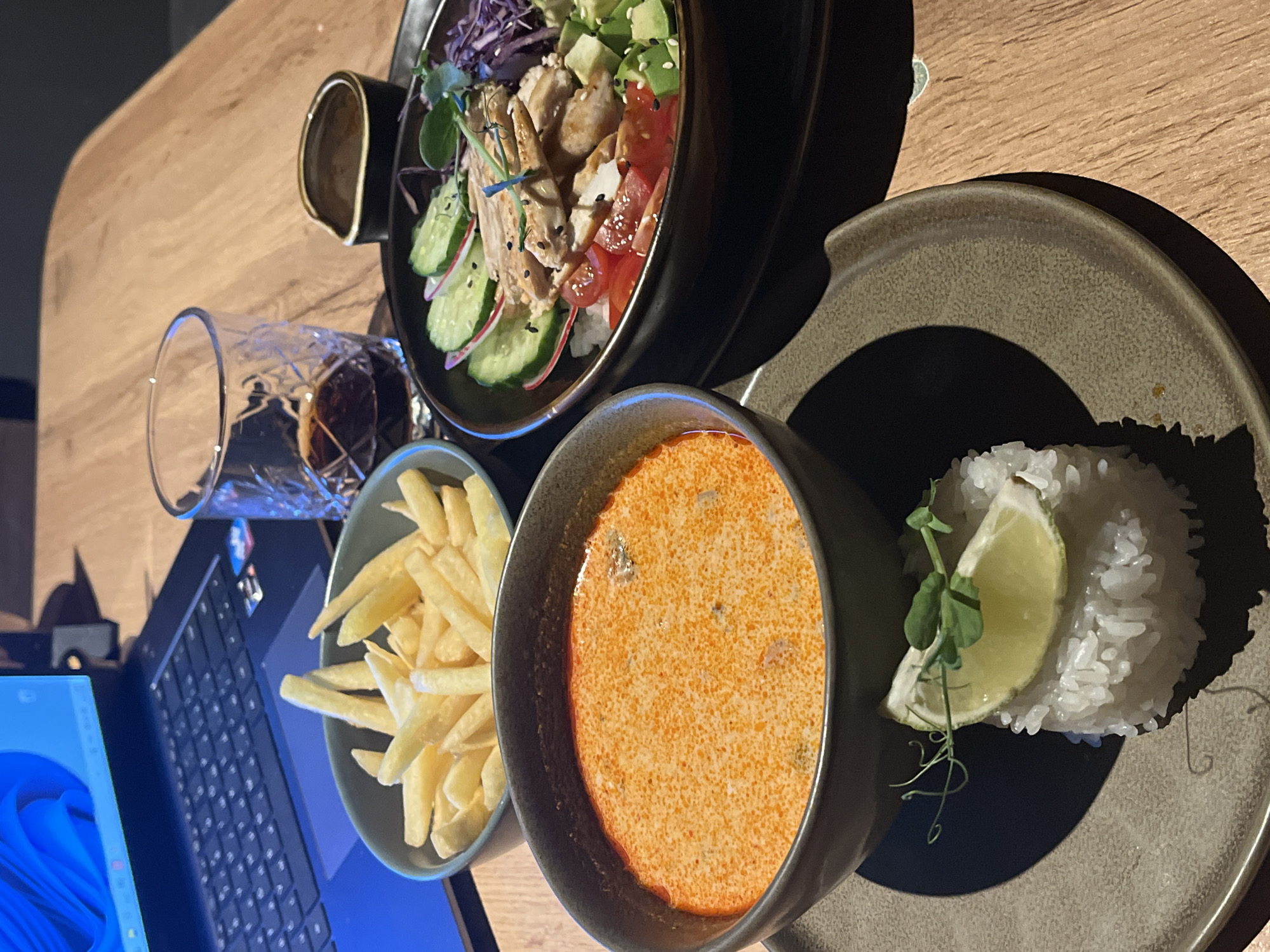 On a wooden table in Unknown, Unknown, a variety of dishes are arranged near a laptop and a glass of liquid. At the top, there is a dark bowl containing a salad with chicken, avocado, red cabbage, cucumbers, and tomatoes over a base of rice. Beside it, a brown plate holds a bowl of orange soup accompanied by a mound of white rice garnished with a lime wedge and sprouts. Adjacent to these dishes is a bowl filled with french fries. A glass with a dark liquid is near the fries and a partially visible blue laptop sits at the edge of the frame.