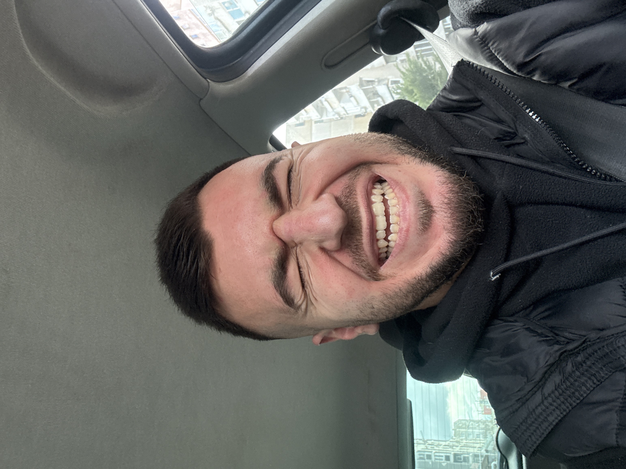A medium shot, taken from a high angle in the front seat of a vehicle, depicts a man with dark hair and a beard wearing a black hooded jacket, who is either grimacing or laughing, exposing his teeth. The interior of the vehicle is visible in the background with the headliner in the top left, and the window frame visible as well. Outside the window in the upper portion of the photo, a building is seen that suggests that the location is likely in a city. While it is difficult to ascertain a specific location without additional visual cues, the presence of modern architecture in the background is consistent with the urban landscape of Sofia, Bulgaria, though this is not definitive.